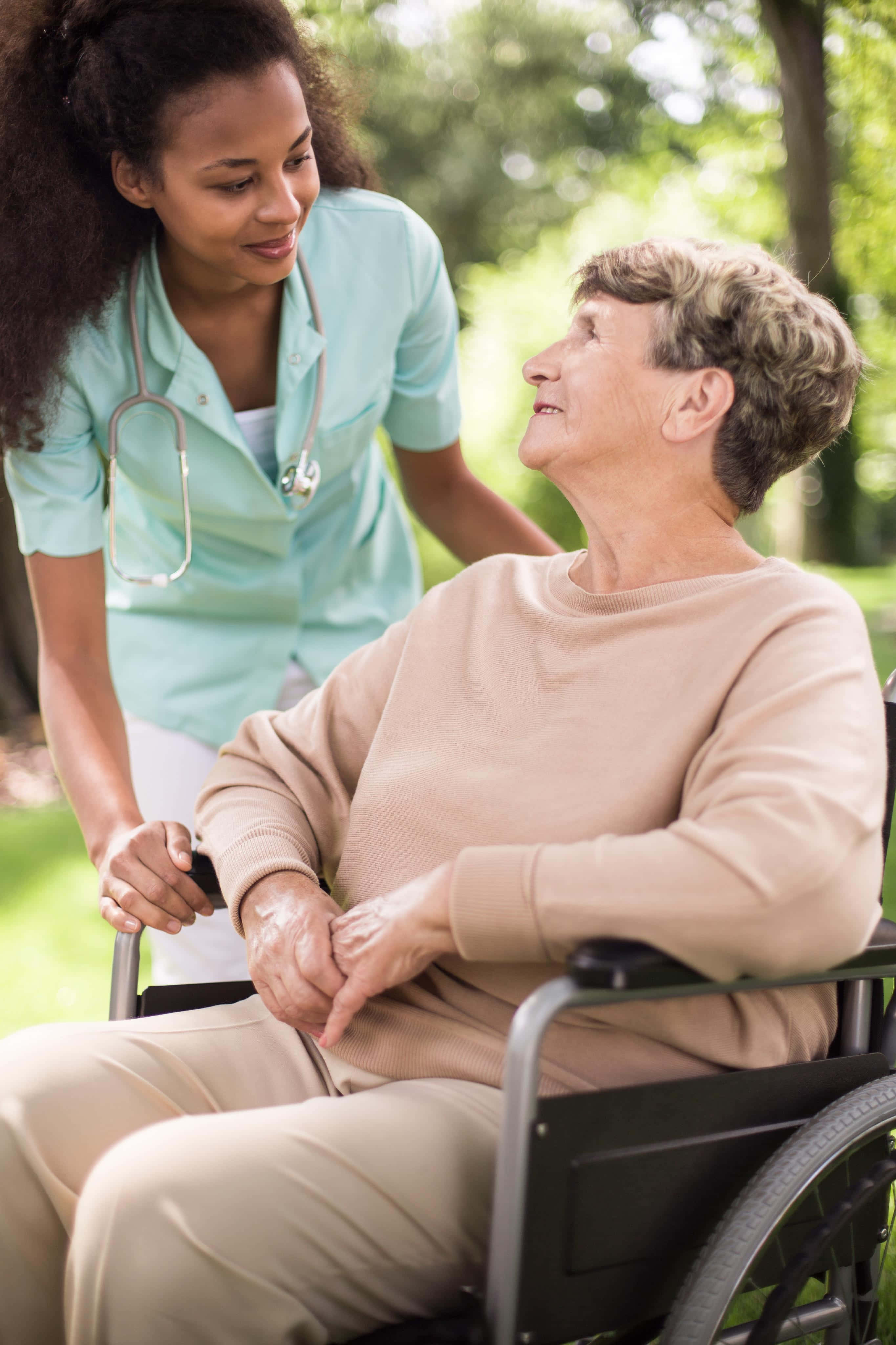 Nursing 桌布 壁紙 — 一位身穿青綠色制服嘅年輕護士，喺陽光普照、綠樹成蔭嘅公園入面，輕輕噉幫助一位坐輪椅嘅老婦人。