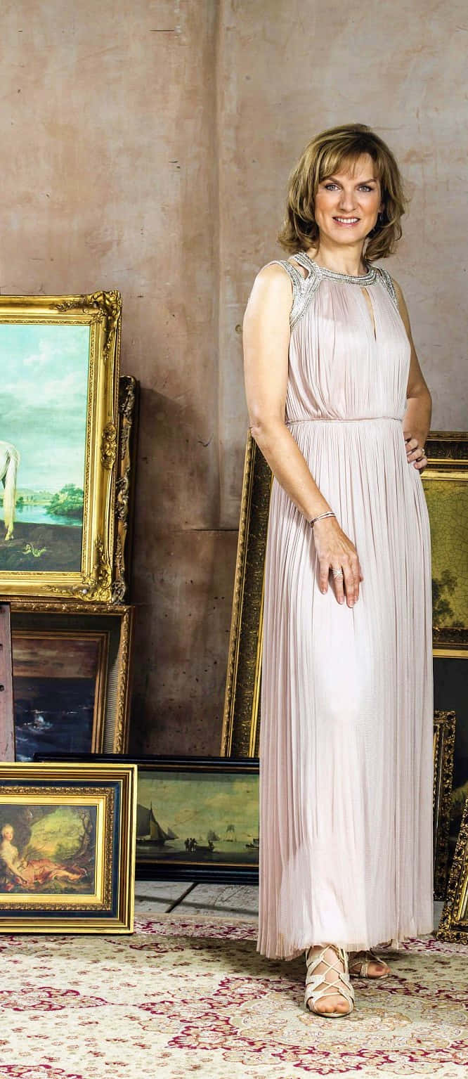 Fiona Bruce 桌布 壁紙 — 一位身穿飄逸淡粉紅色長裙的女性，在溫暖燈光的畫廊中，於華麗的金框畫作間優雅微笑。