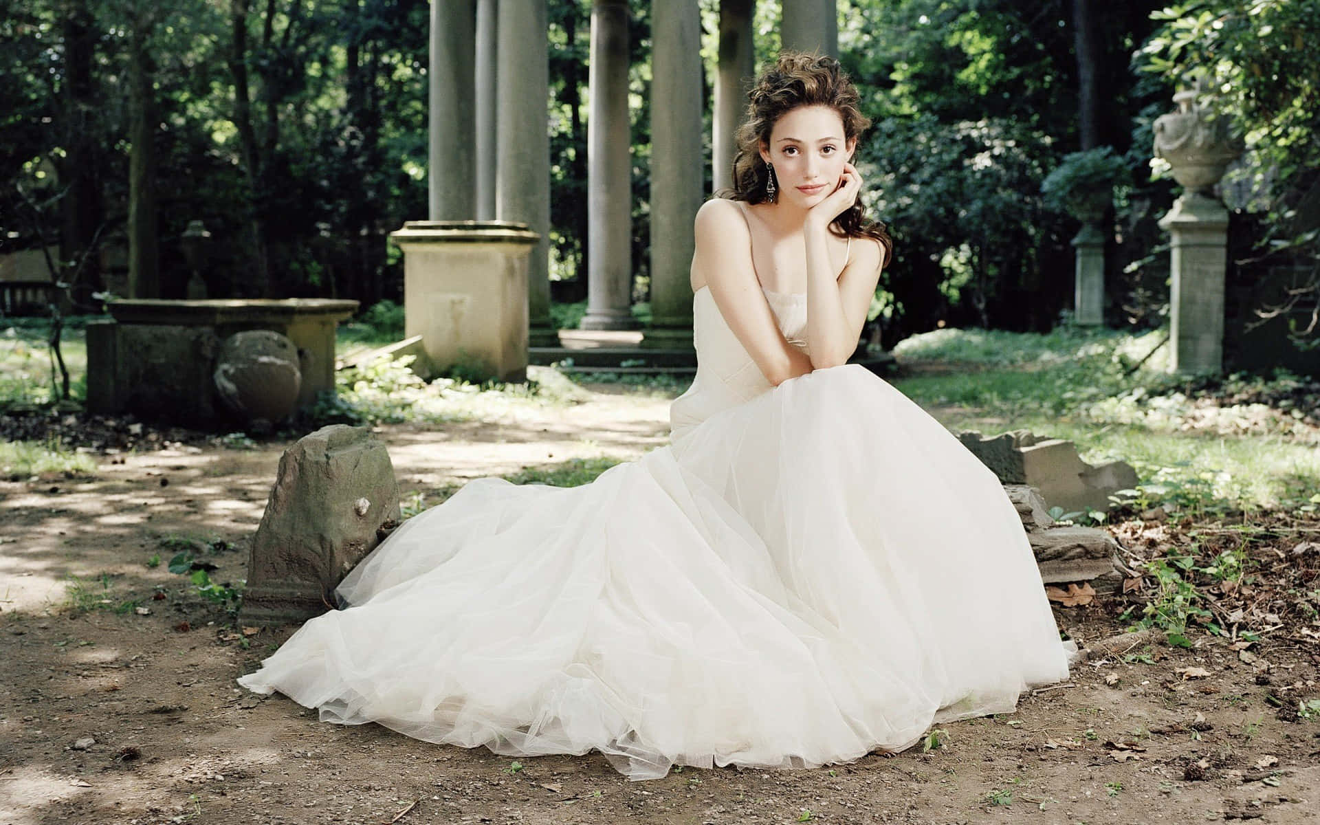 Wedding Dress 桌布 壁紙 — 一位身穿飄逸白色婚紗的女子，優雅地坐在陽光斑駁的森林中的古老石頭廢墟中。