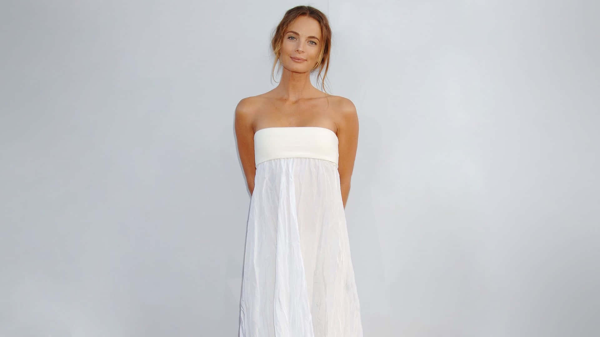 Gabrielle Anwar 桌布 壁紙 — 一位身穿飄逸白色無肩帶連衣裙的女性在柔和灰色工作室背景前優雅站立，光線柔和。
