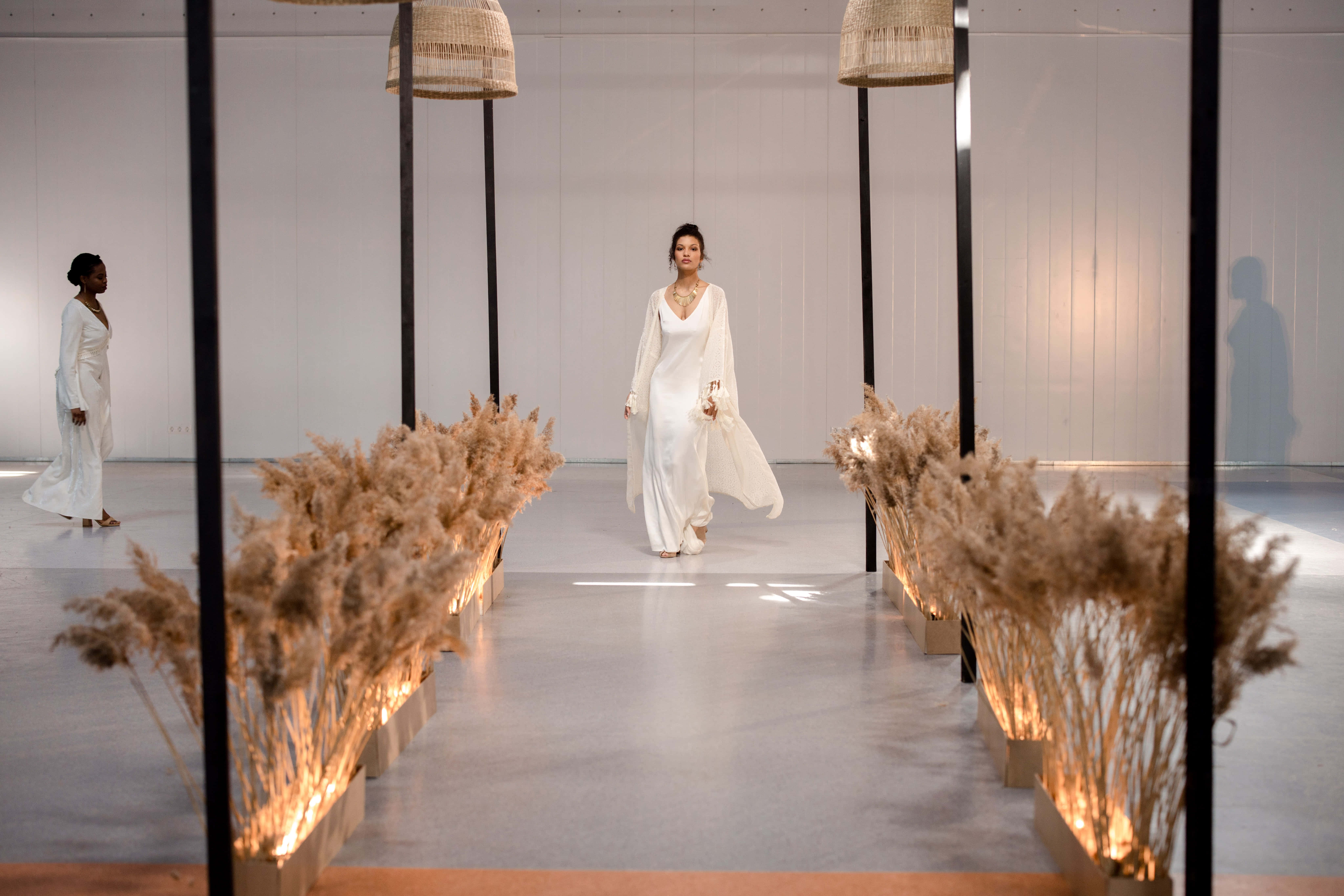 Runway 桌布 壁紙 — 一位身穿飄逸白色長裙的女性，在兩旁佈滿發光蒲葦裝飾的簡約天橋上行走。