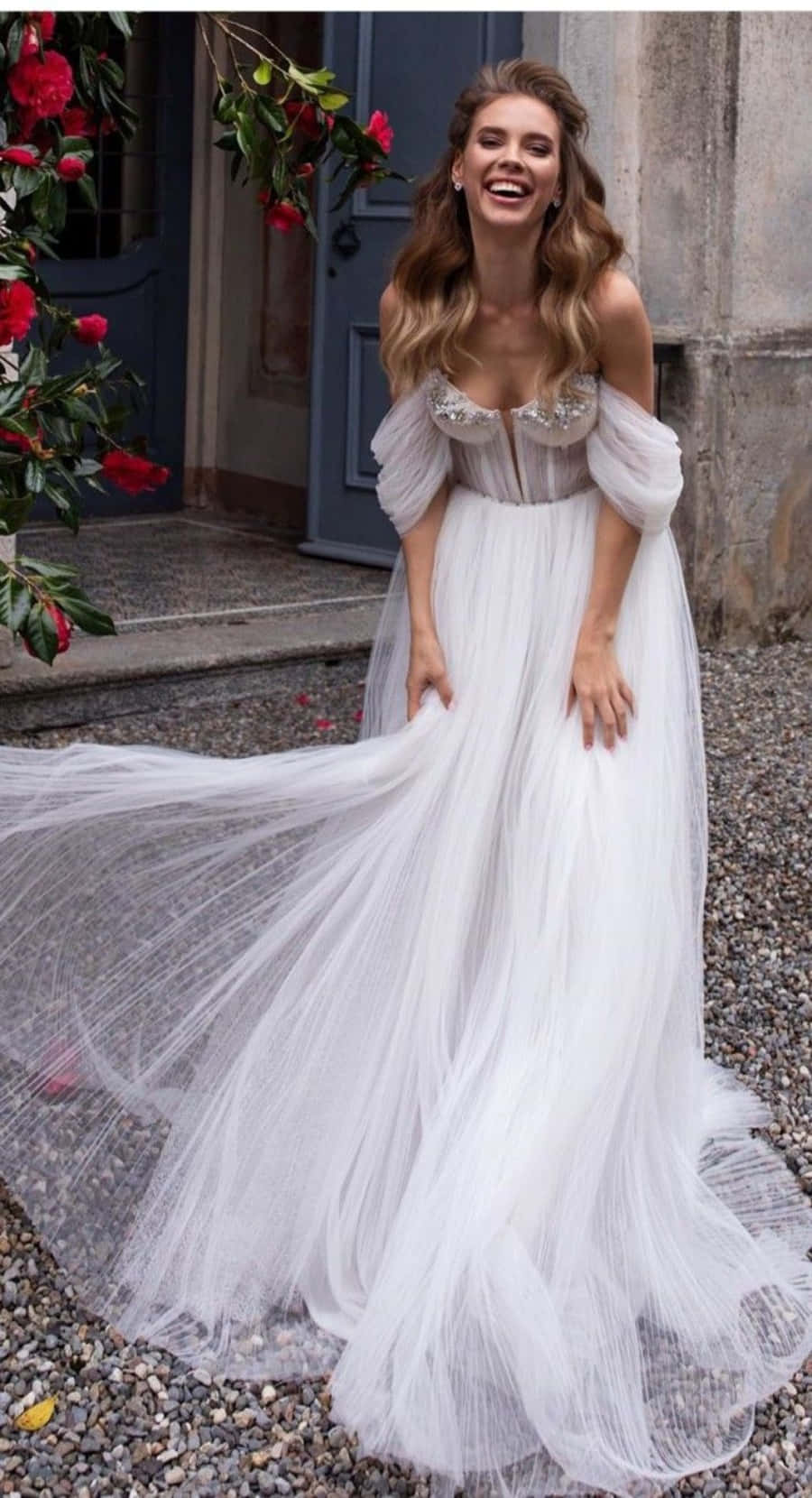 Wedding Dress 桌布 壁紙 — 一位身穿飄逸白色長裙的快樂女士在戶外藍色門和紅花旁邊大笑。