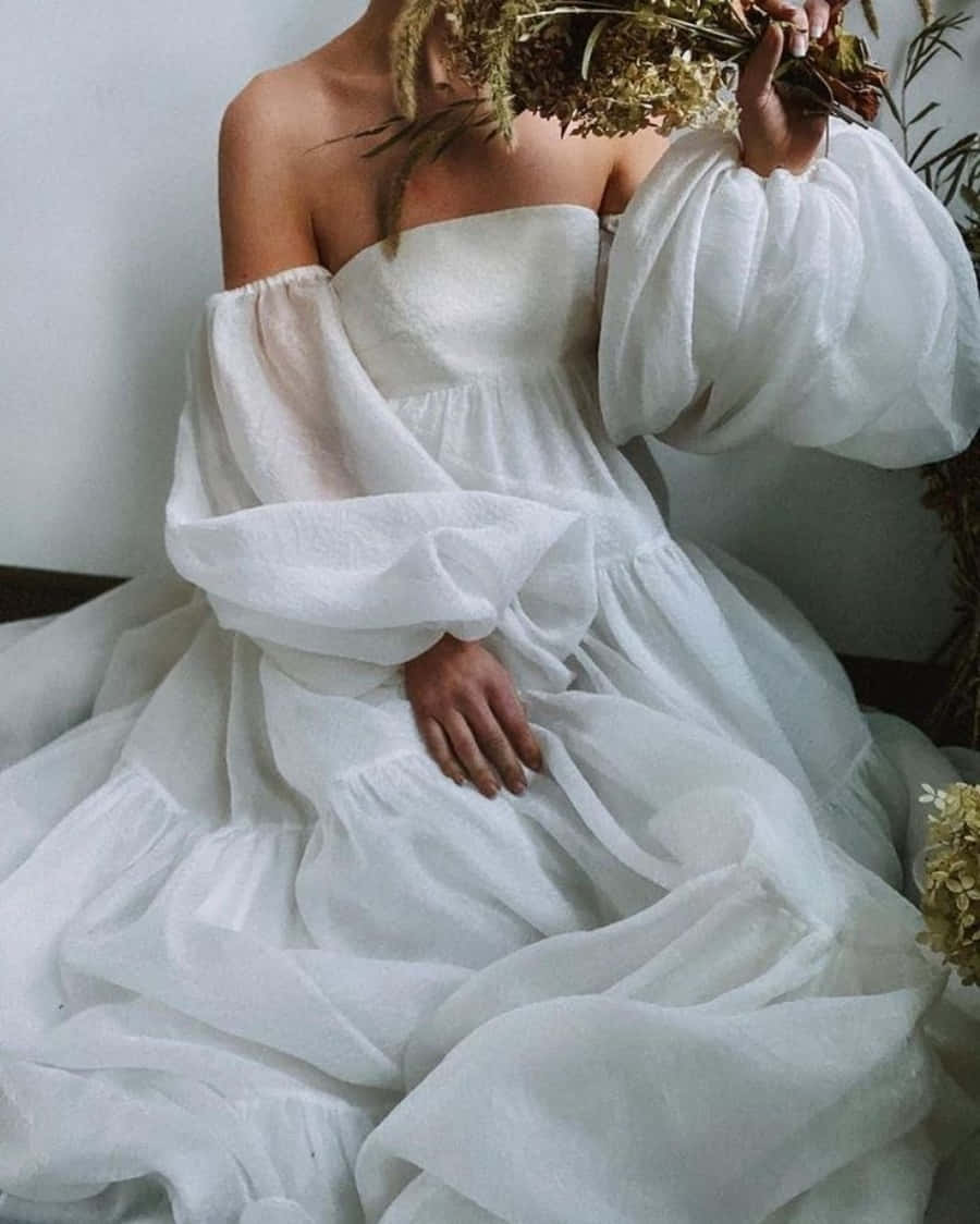 Wedding Dress 桌布 壁紙 — 一位身穿飄逸白色露肩長裙的女性，在素牆前將乾花舉到臉龐。