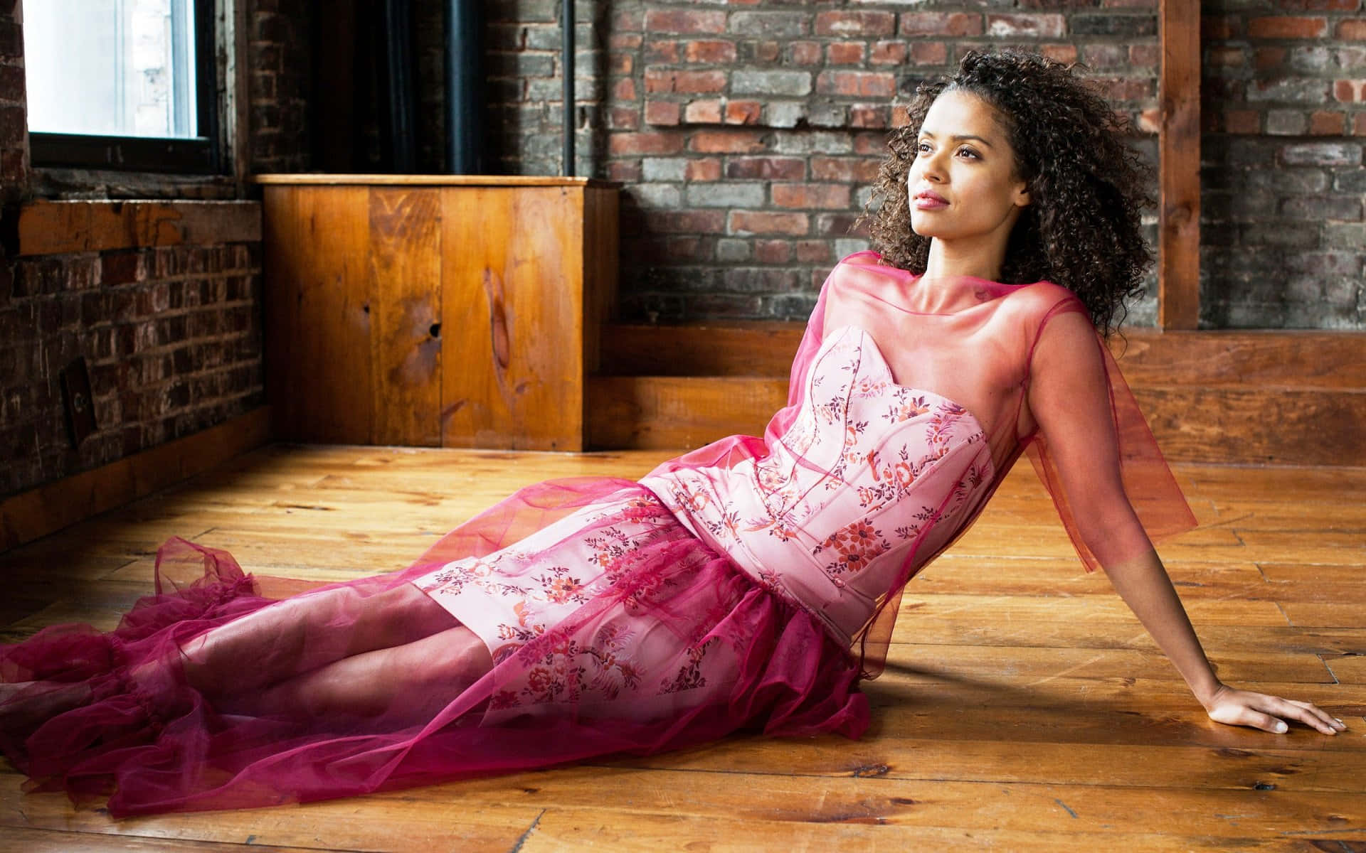Gugu Mbatha-raw 桌布 壁紙 — 一位身穿飄逸粉紅花卉長裙的女性，優雅地斜倚在木地板上，背景是質樸的磚牆。