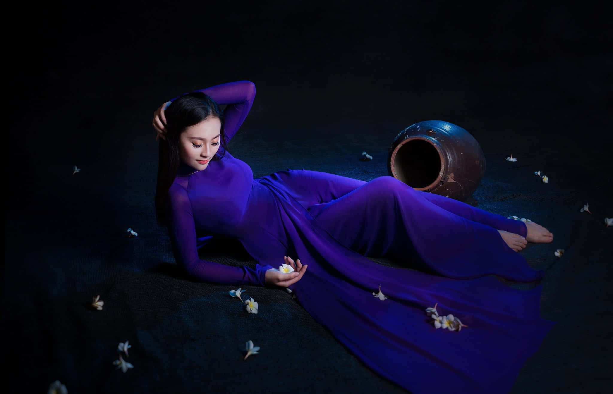 Purple Dress 桌布 壁紙 — 一位身穿飄逸紫色連衣裙的女性優雅地躺在黑暗的地面上，手持一朵白花，旁邊放著一個陶瓷罐。