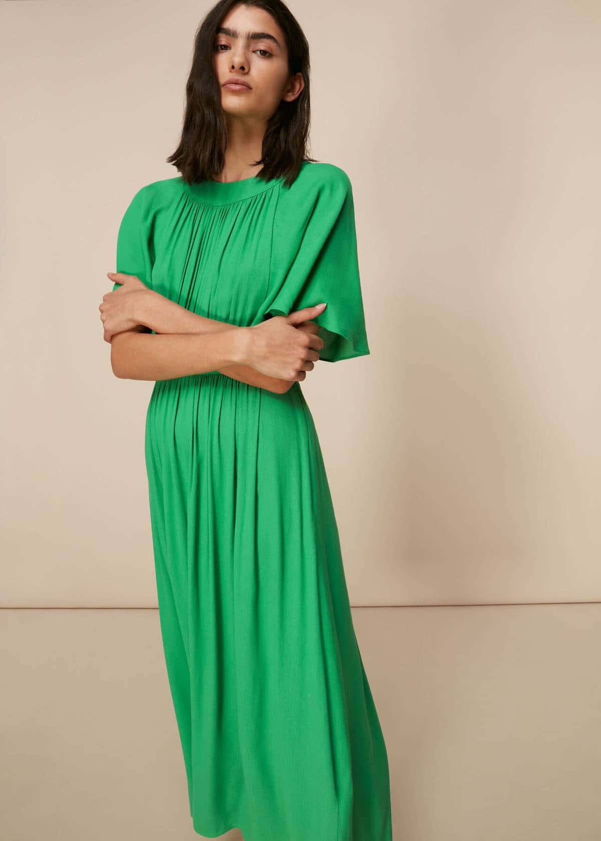 Green Fashion 桌布 壁紙 — 一位身穿飄逸翡翠綠連衣裙的女性，雙臂交叉，靠著中性米色工作室牆壁站立。