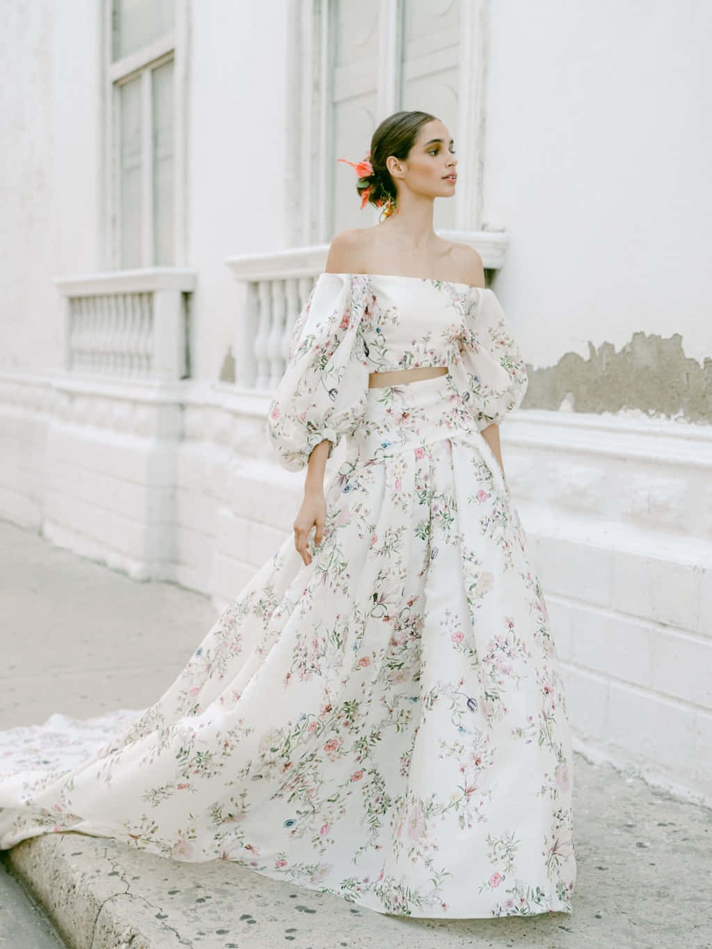 Bride's Dress 桌布 壁紙 — 一位身穿飄逸花卉露肩長裙的女性，優雅地站在一棟風化的白色建築外牆前。