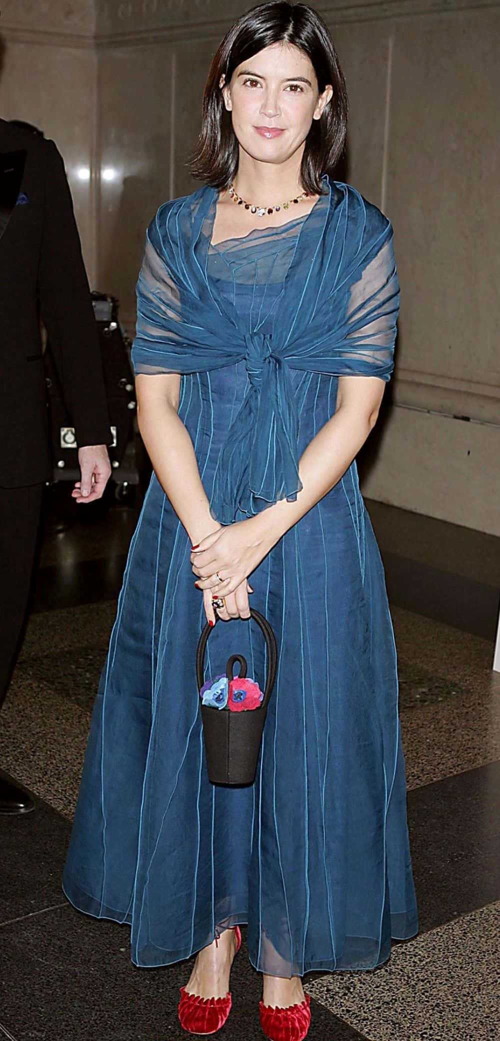 Phoebe Cates 桌布 壁紙 — 一位身穿飄逸藍色連衣裙和紅色鞋子的女士，手持一個帶有彩色花朵的小黑色手袋。
