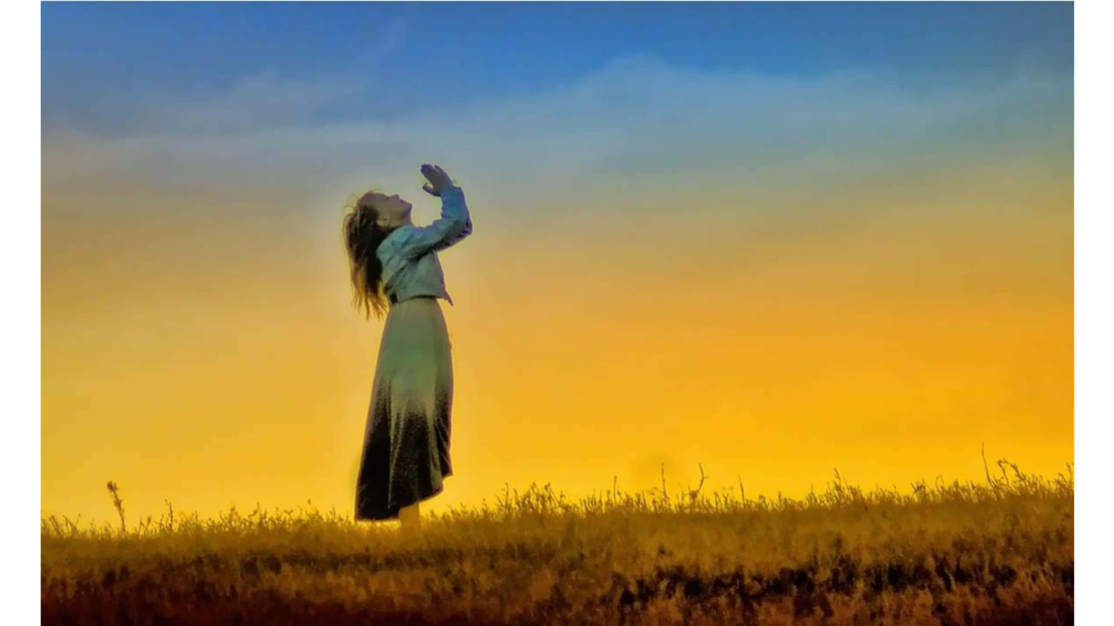 Praying 桌布 壁紙 — 一位身穿飄逸長裙的女子站在日落時分的草地上，雙臂向金色的天空高舉。
