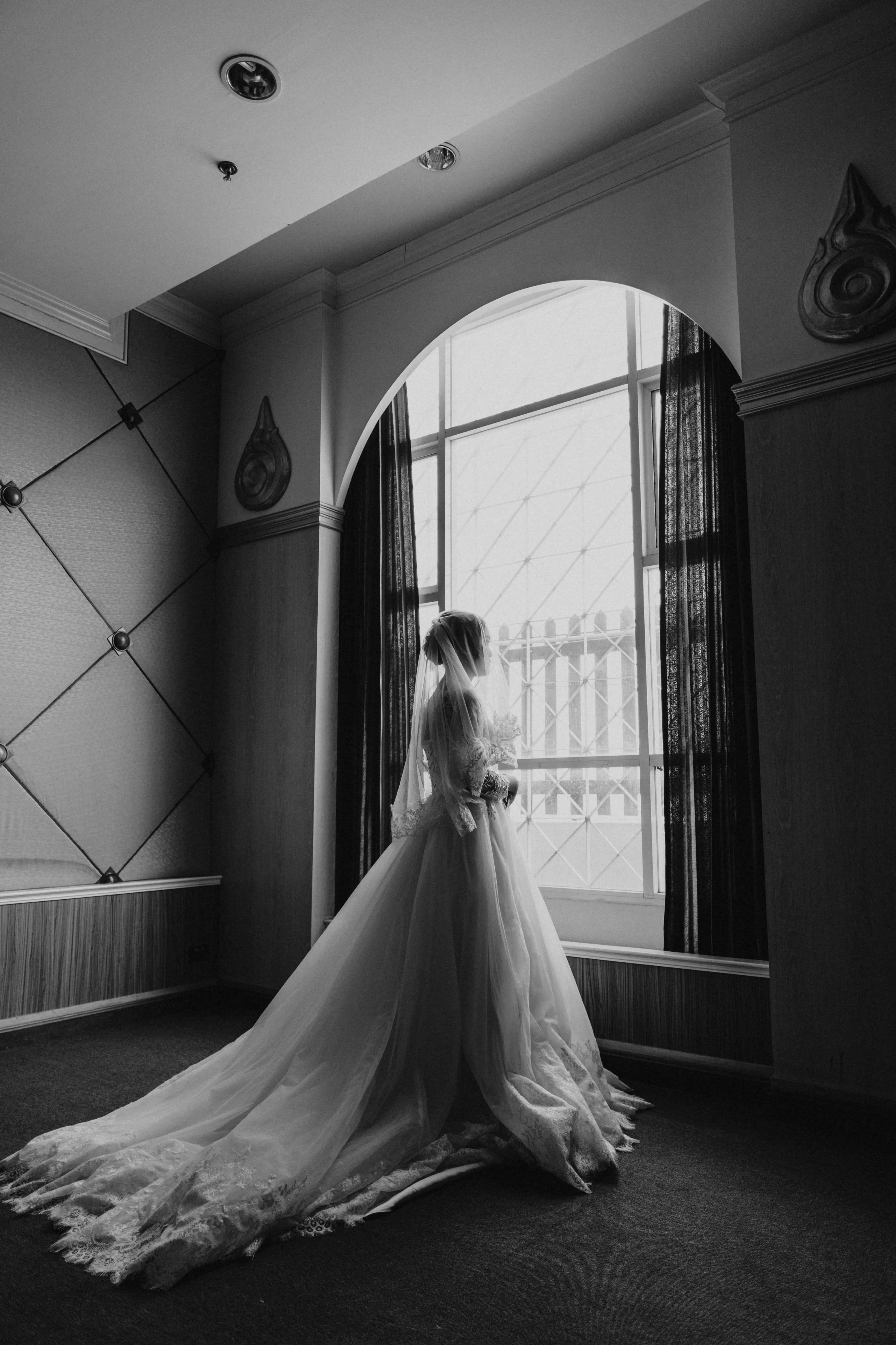 Black And White Wedding 桌布 壁紙 — 一位身穿飄逸長裙的新娘，側身站在拱形窗戶旁，頭紗和裙擺如瀑布般垂下，以單色優雅凝視窗外。