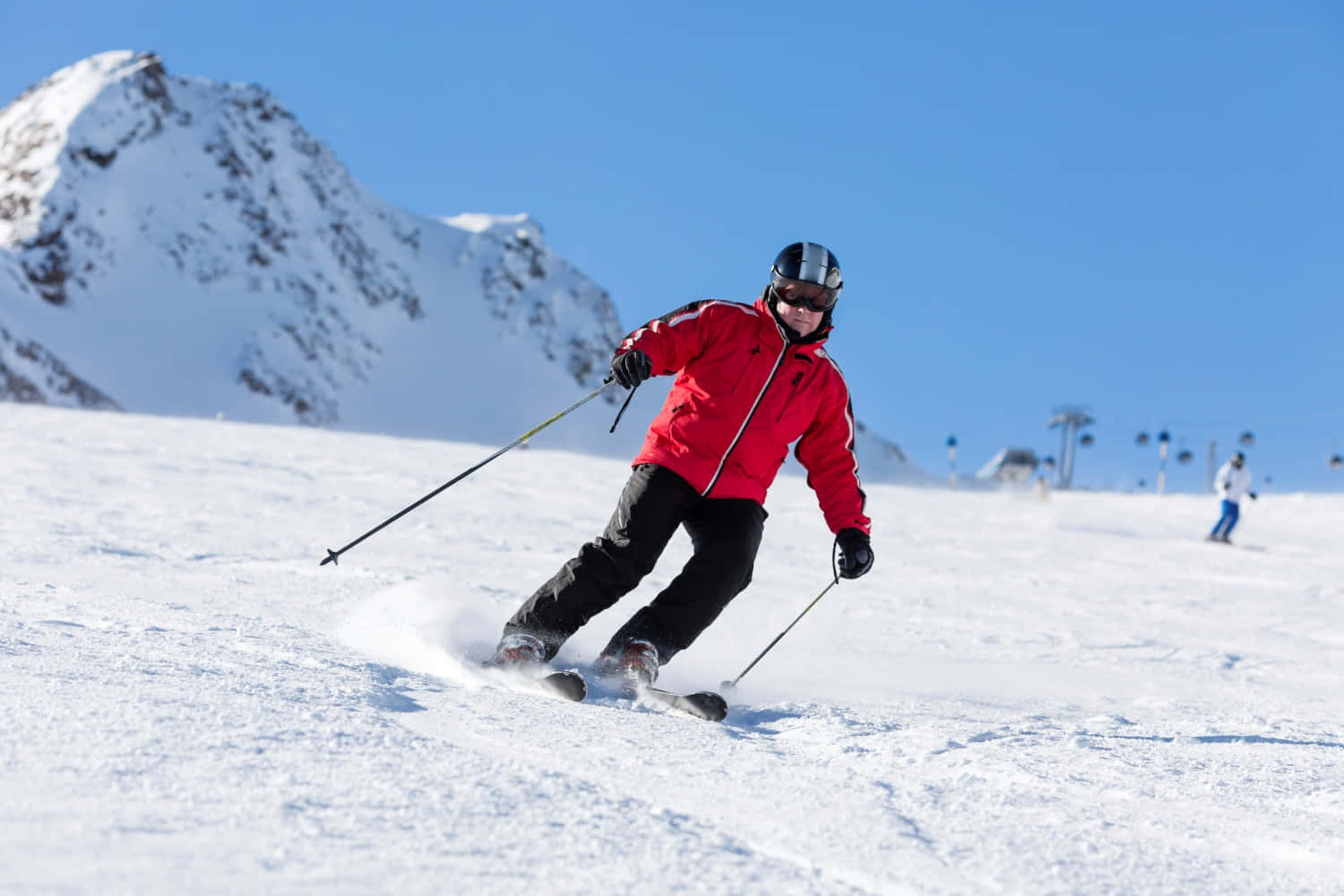 Skiing 桌布 壁紙 — 一位身穿鮮紅色外套的滑雪者正在白雪皚皚的斜坡上滑行，身後雪花飛濺，背景是白雪覆蓋的山峰和清澈的藍天。