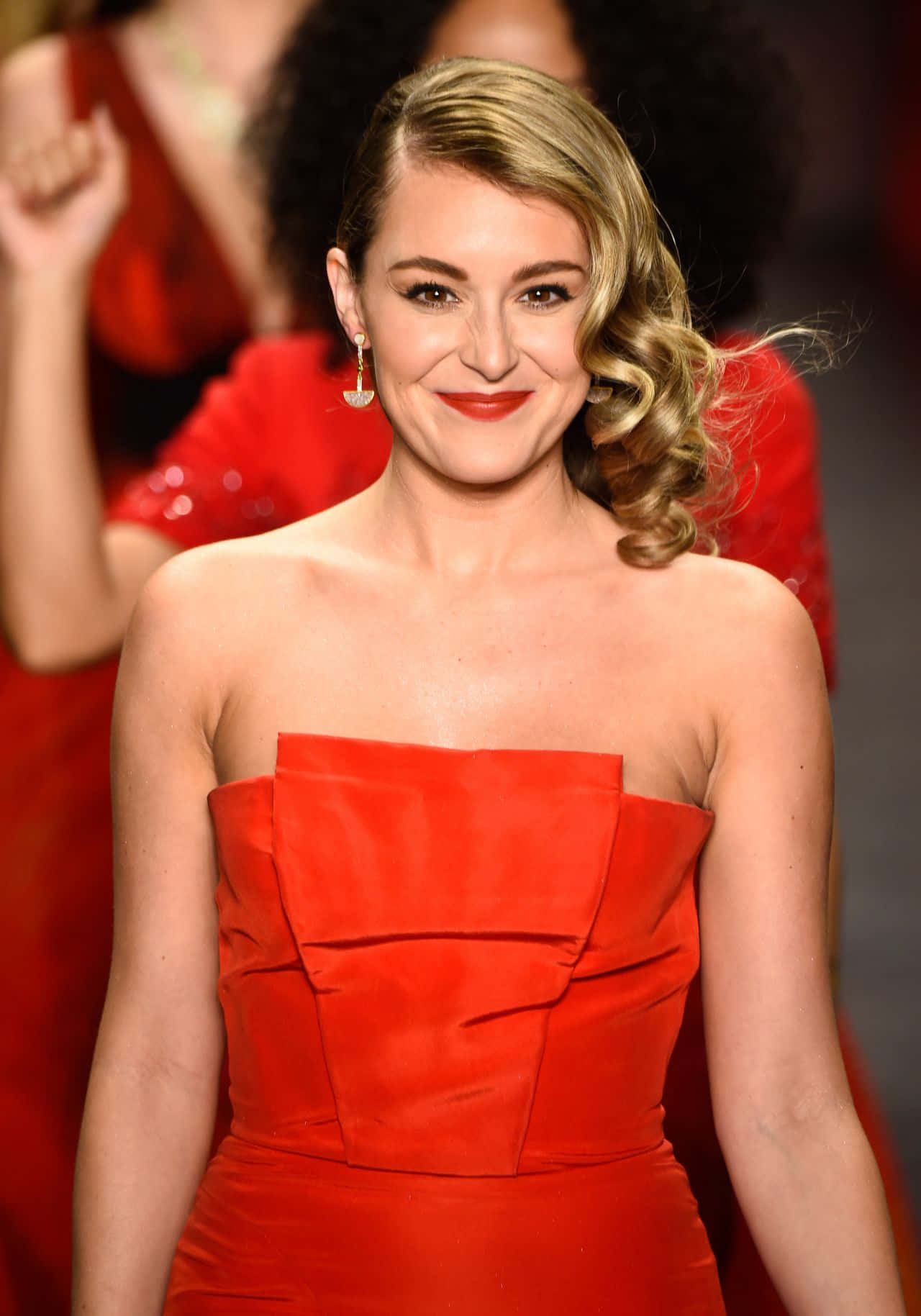 Alexa Penavega 桌布 壁紙 — 一位身穿鮮紅色無肩帶禮服的女士在時裝天橋上自信地微笑，身後有模糊的紅色模特兒。