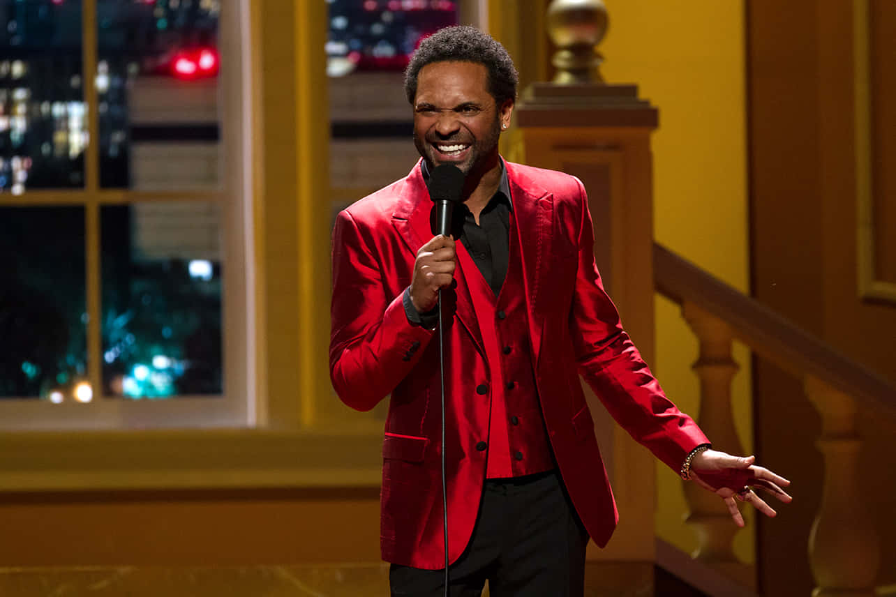 Mike Epps 桌布 壁紙 — 一位身穿鮮紅色西裝的喜劇演員在溫暖燈光的舞台上，手持麥克風開懷大笑，身後窗戶可見城市景觀。