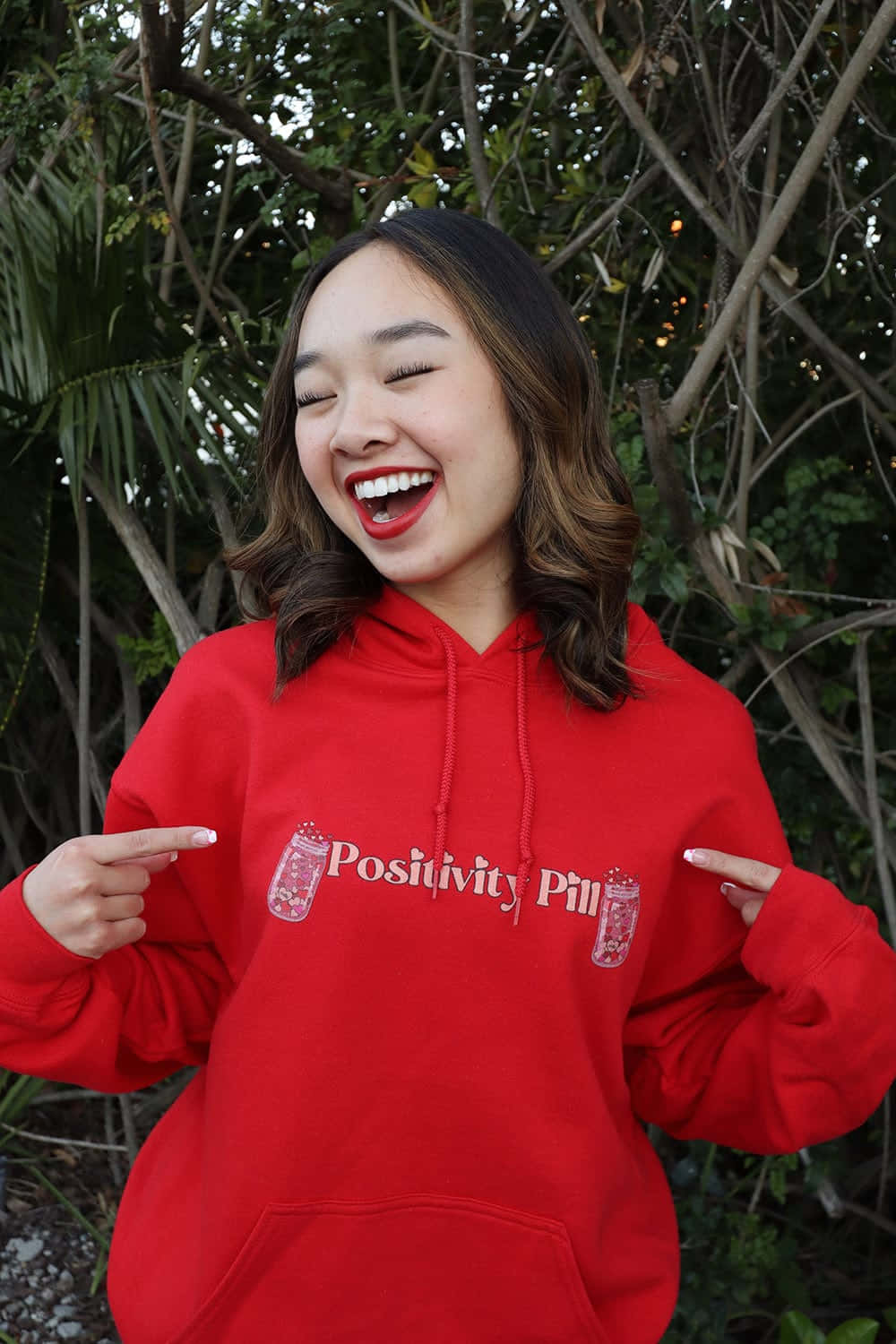 Red Hoodie 桌布 壁紙 — 一位身穿鮮紅色連帽衫的快樂年輕女性，笑著指向胸前「Positivity Pill」圖案。