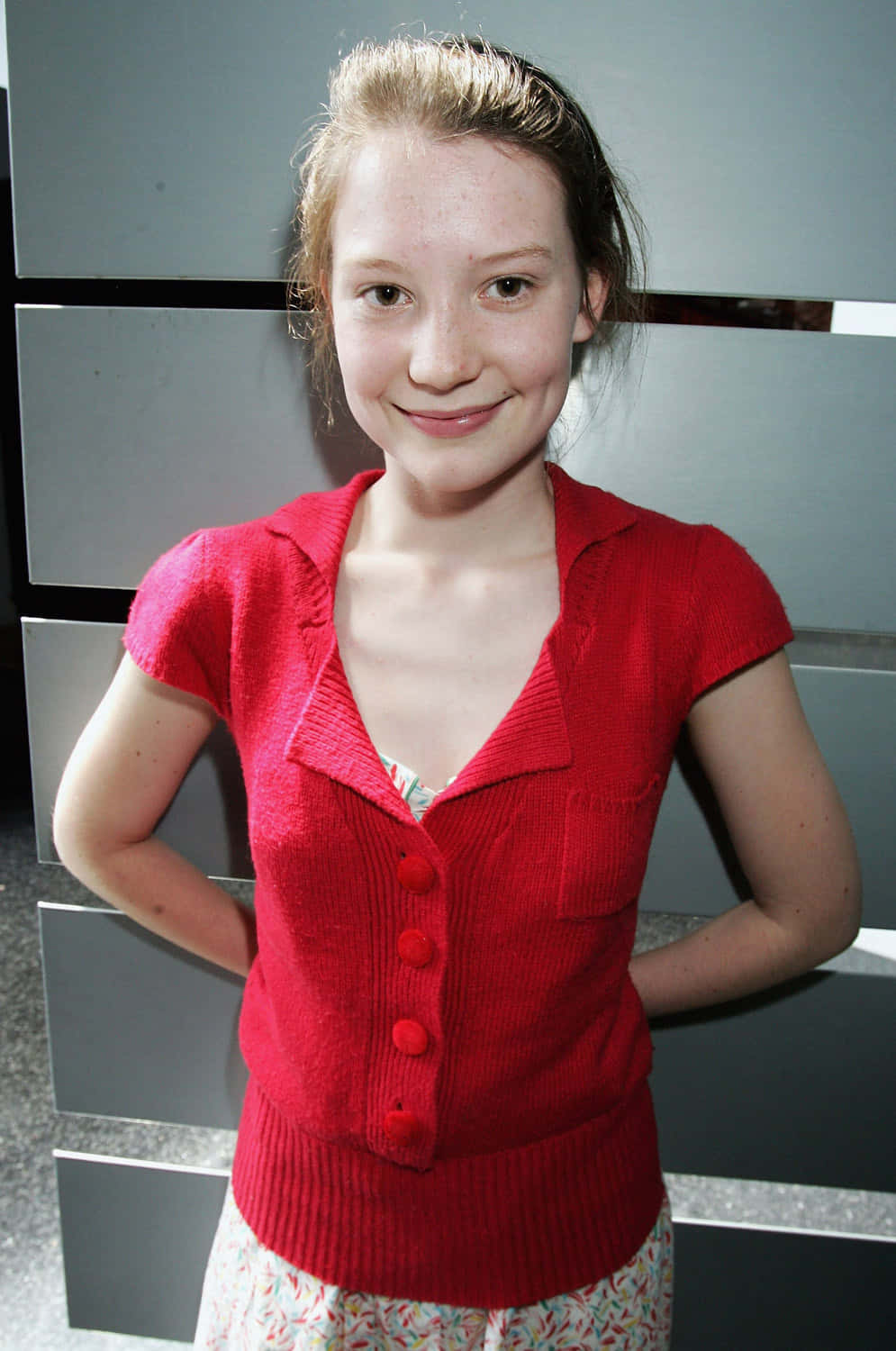 Mia Wasikowska 桌布 壁紙 — 一位身穿鮮紅色針織開衫的年輕女孩，在現代灰色鑲板牆前溫柔地微笑。