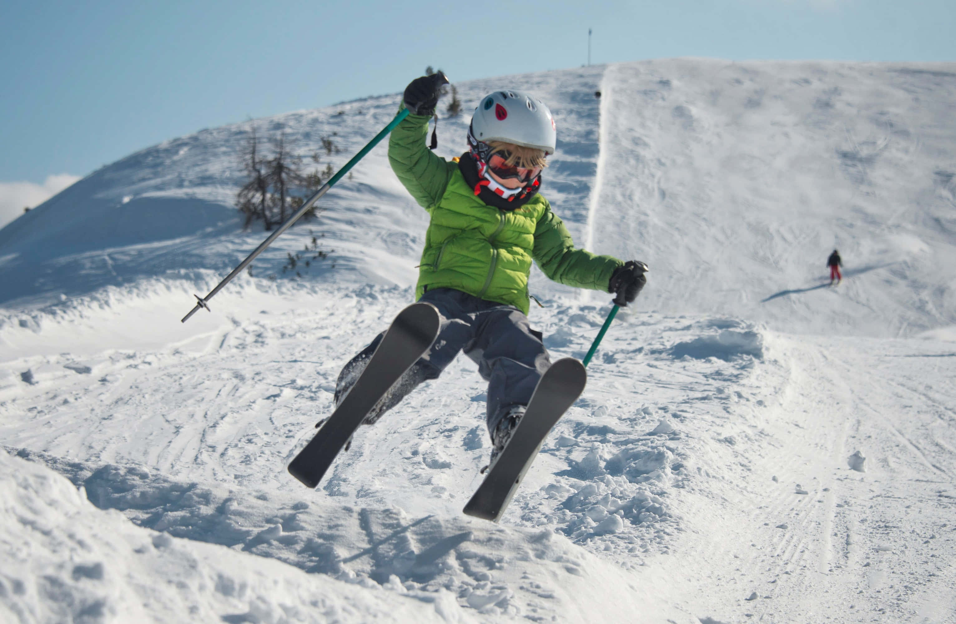 Skiing 桌布 壁紙 — 一位身穿鮮綠色外套的年輕滑雪者，在晴朗的藍天下，飛越白雪覆蓋的山坡。