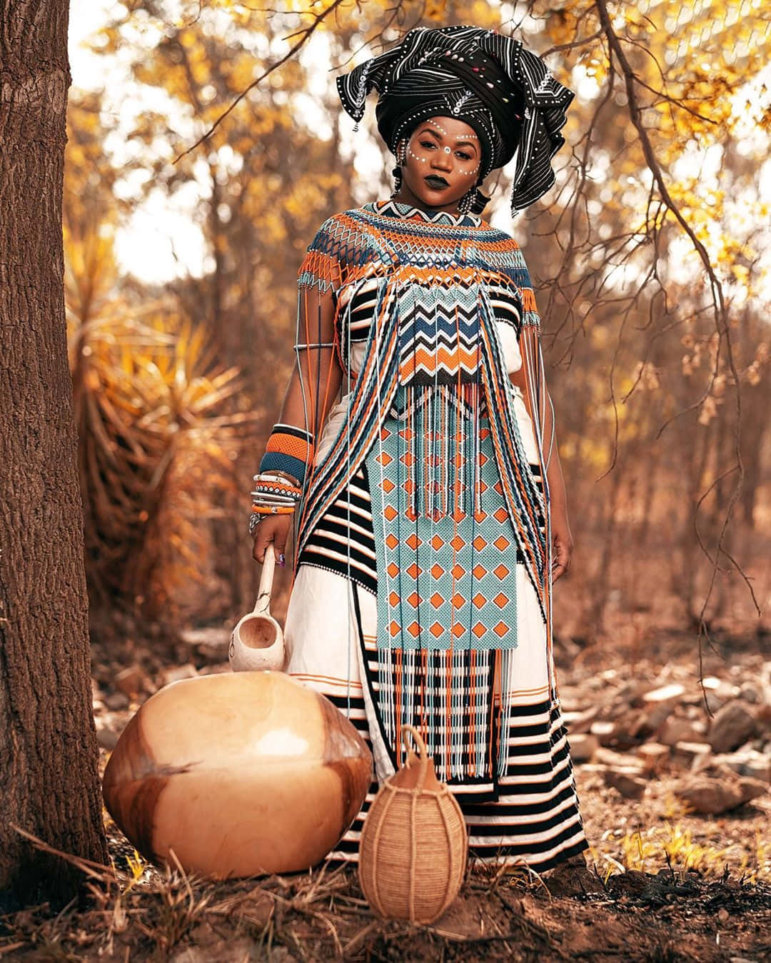African Culture 桌布 壁紙 — 一位身穿鮮豔幾何圖案傳統服飾的女性，站在陽光明媚的森林中，手持葫蘆和籃子。