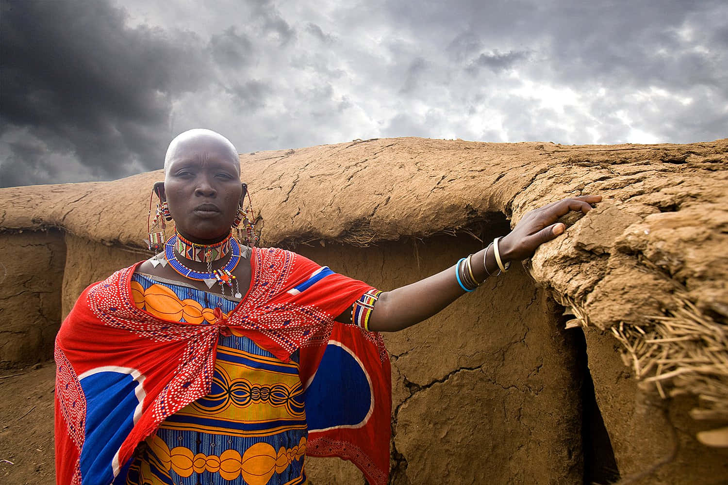 African Culture 桌布 壁紙 — 一位身穿鮮豔紅、藍、橙色傳統服飾的馬賽族婦女，站在風暴天空下的泥磚房屋前。