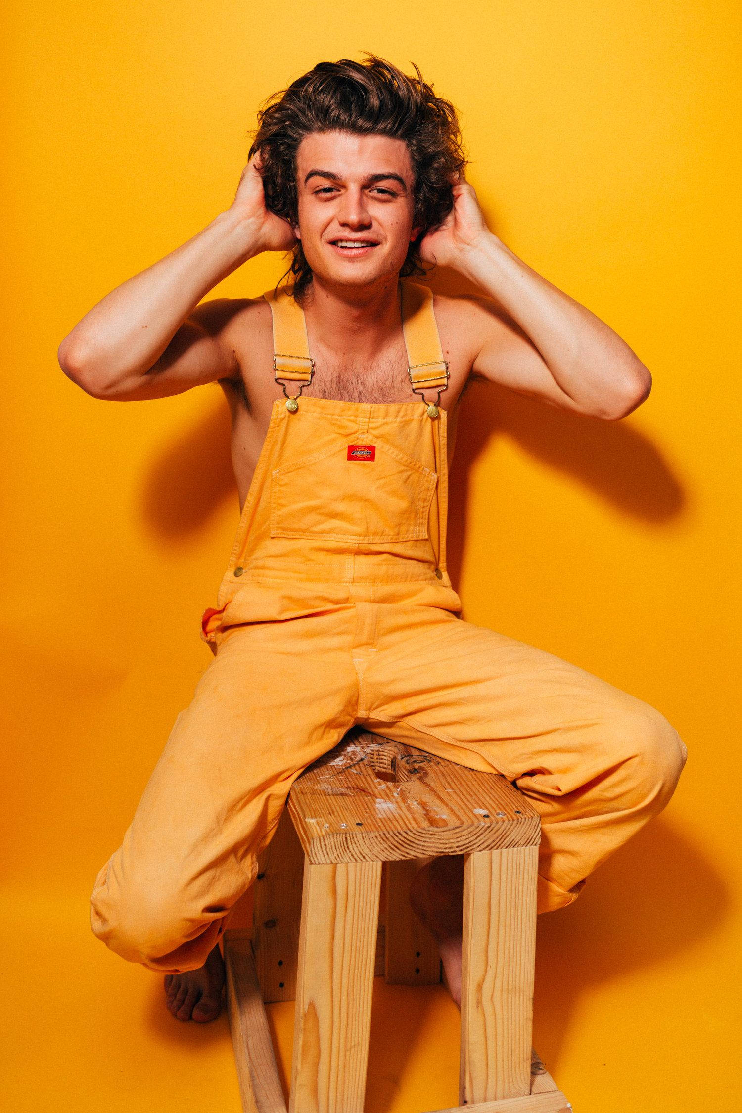 Joe Keery 桌布 壁紙 — 一位身穿黃色工裝褲的年輕男子坐在木凳上，背景是鮮黃色的牆壁，雙手放在頭上，自信地微笑。