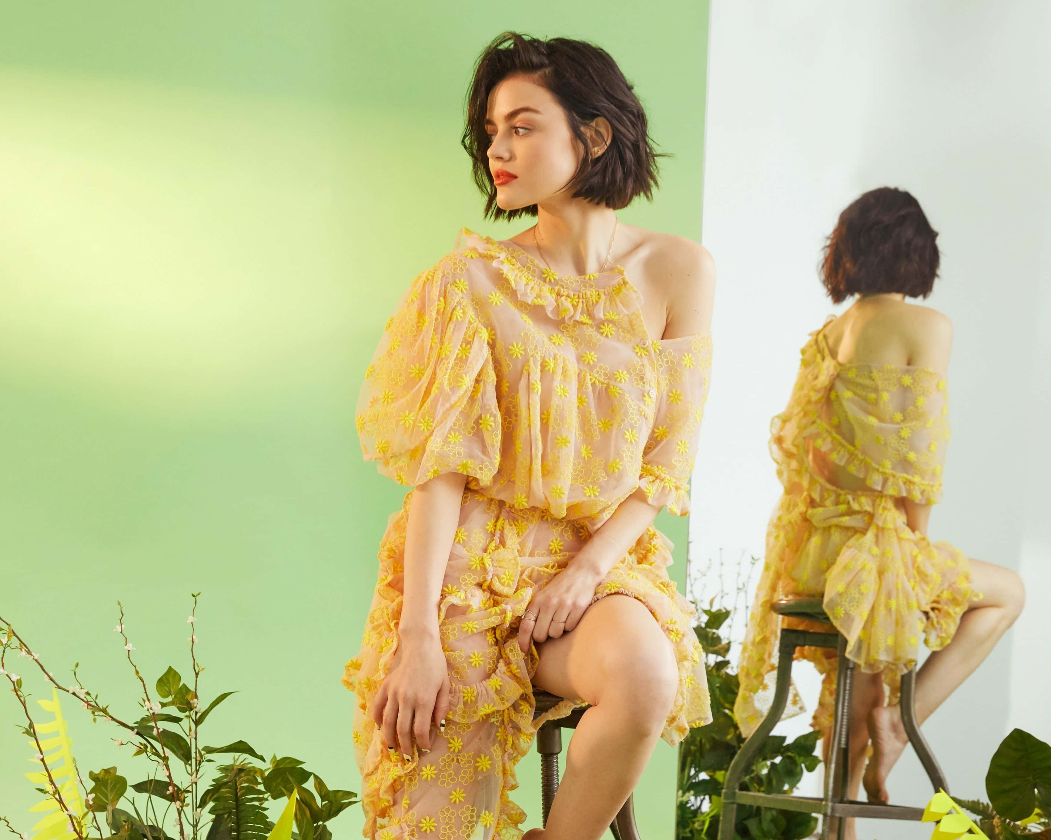 Yellow Dress 桌布 壁紙 — 一位身穿黃色花卉連衣裙的女性坐在凳子上，背景是柔和的綠色牆壁，有植物和鏡子反射。