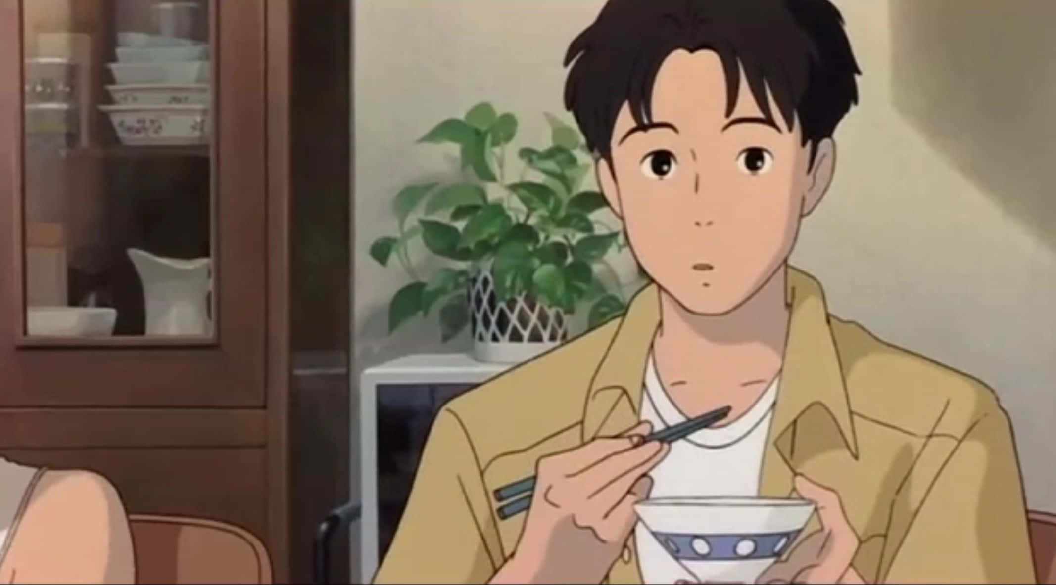 Studio Ghibli Ocean Waves 桌布 壁紙 — 一位身穿黃色襯衫的年輕男子在溫暖燈光的家居環境中用筷子從藍白碗中進食。