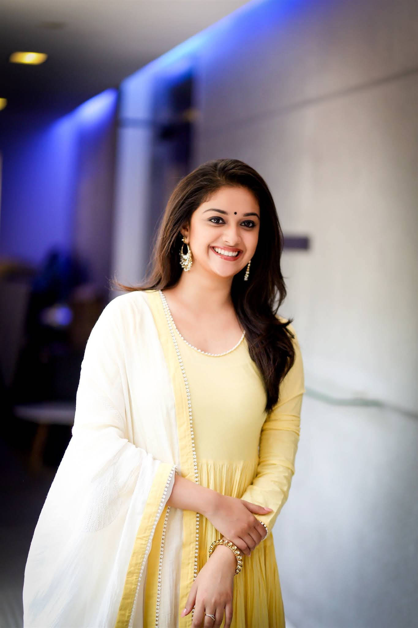 Tamil Actors HD 桌布 壁紙 — 一位身穿黃色連衣裙和白色披肩的容光煥發的女性，在柔和燈光、帶有藍色重點照明的現代室內環境中溫暖地微笑。