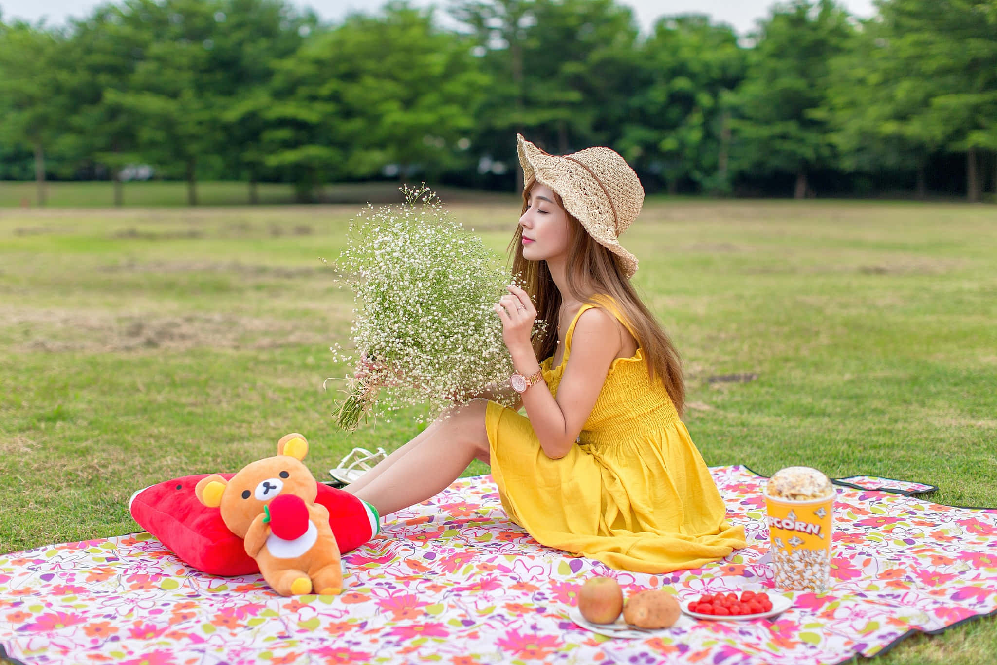 Yellow Dress 桌布 壁紙 — 一位身穿黃色連衣裙和草帽的年輕女子坐在公園草地上的花卉毯子上，手持滿天星花。