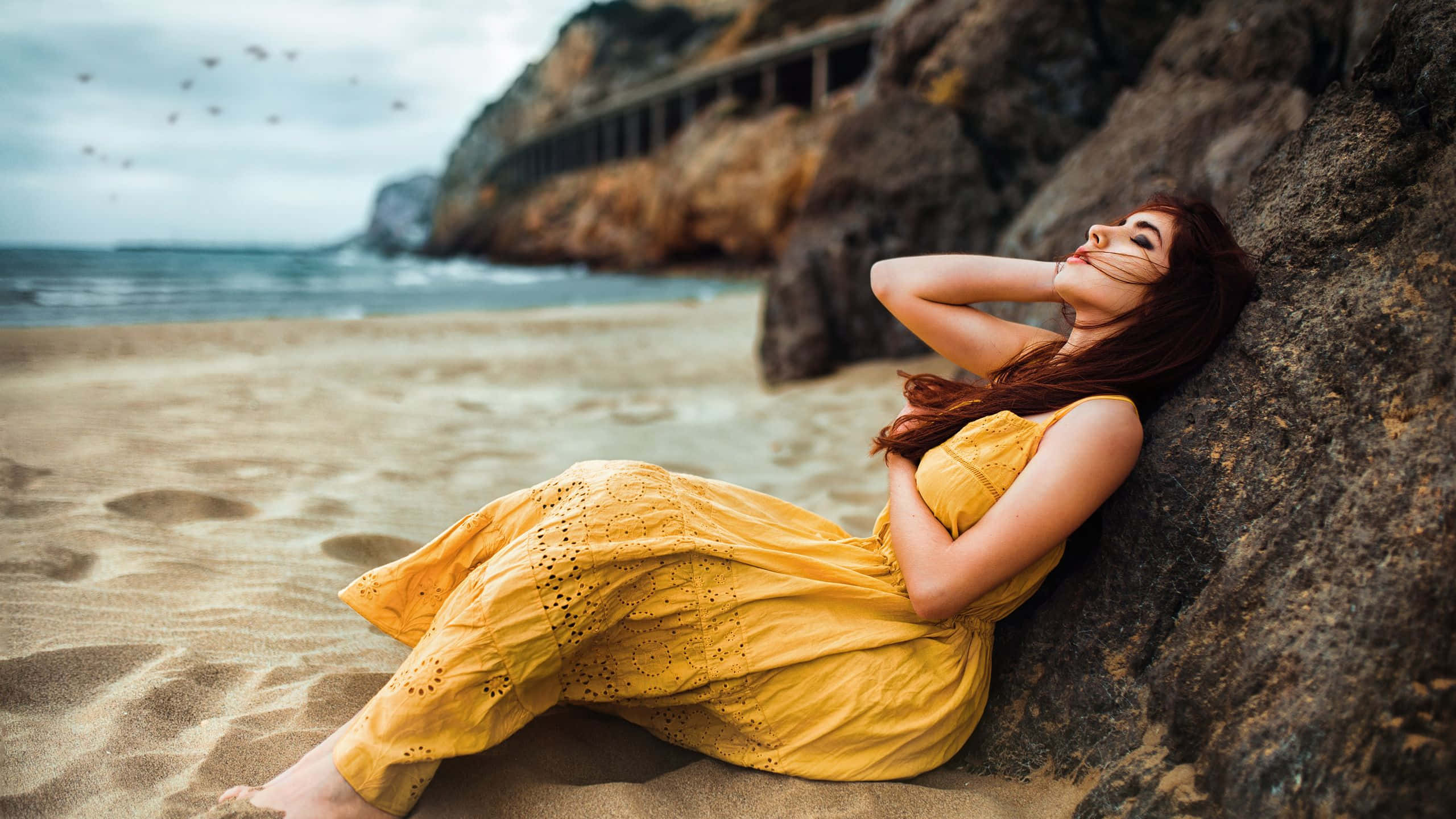 Relaxing Desktop 桌布 壁紙 — 一位身穿黃色連衣裙的女子靠在沙灘上的岩石上，鳥兒在頭頂飛翔，海浪在附近拍打。