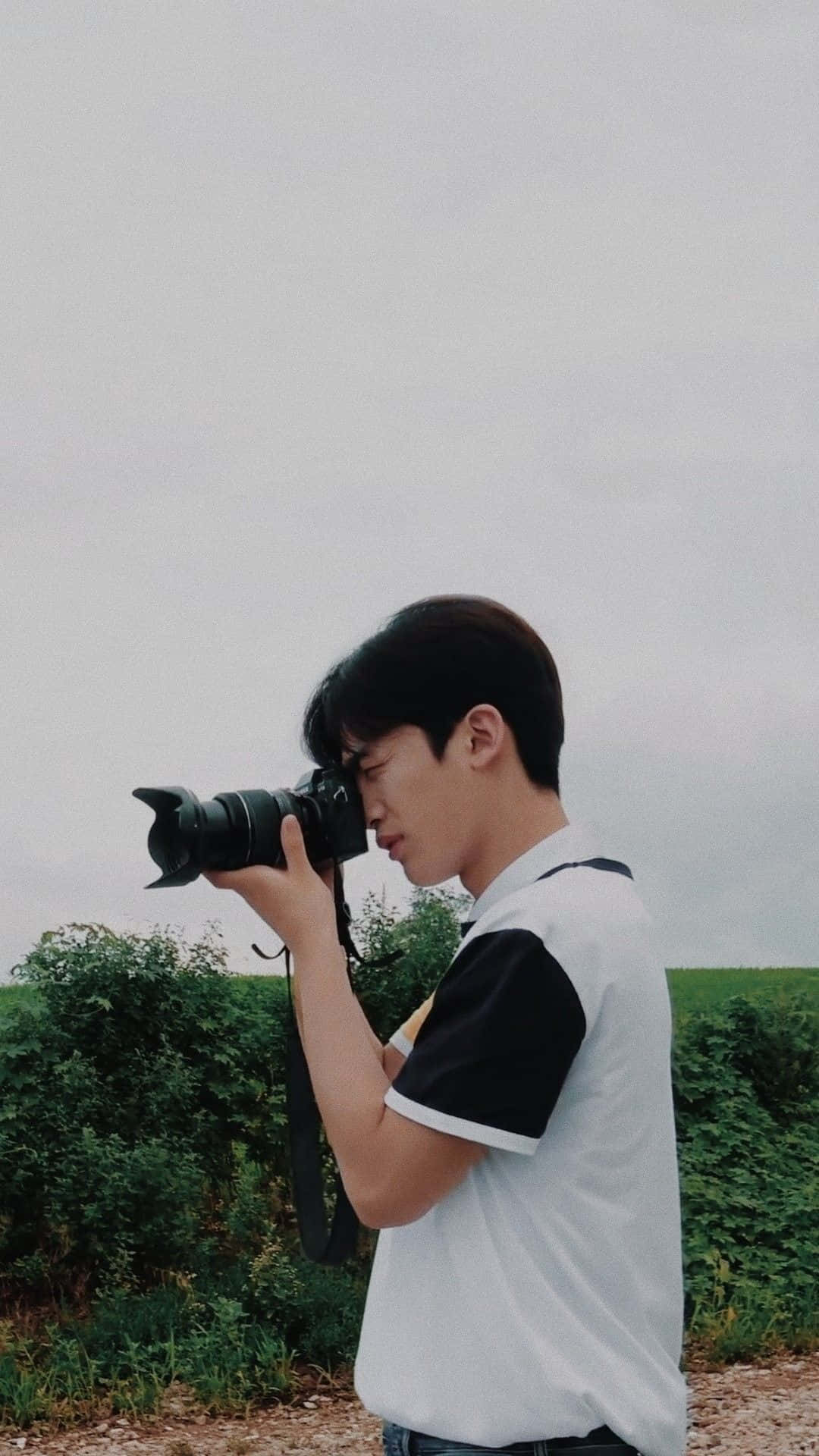 Cute Boyfriend 桌布 壁紙 — 一位身穿黑白襯衫的年輕男子，在陰天和綠色樹葉的背景下，用單反相機捕捉瞬間。