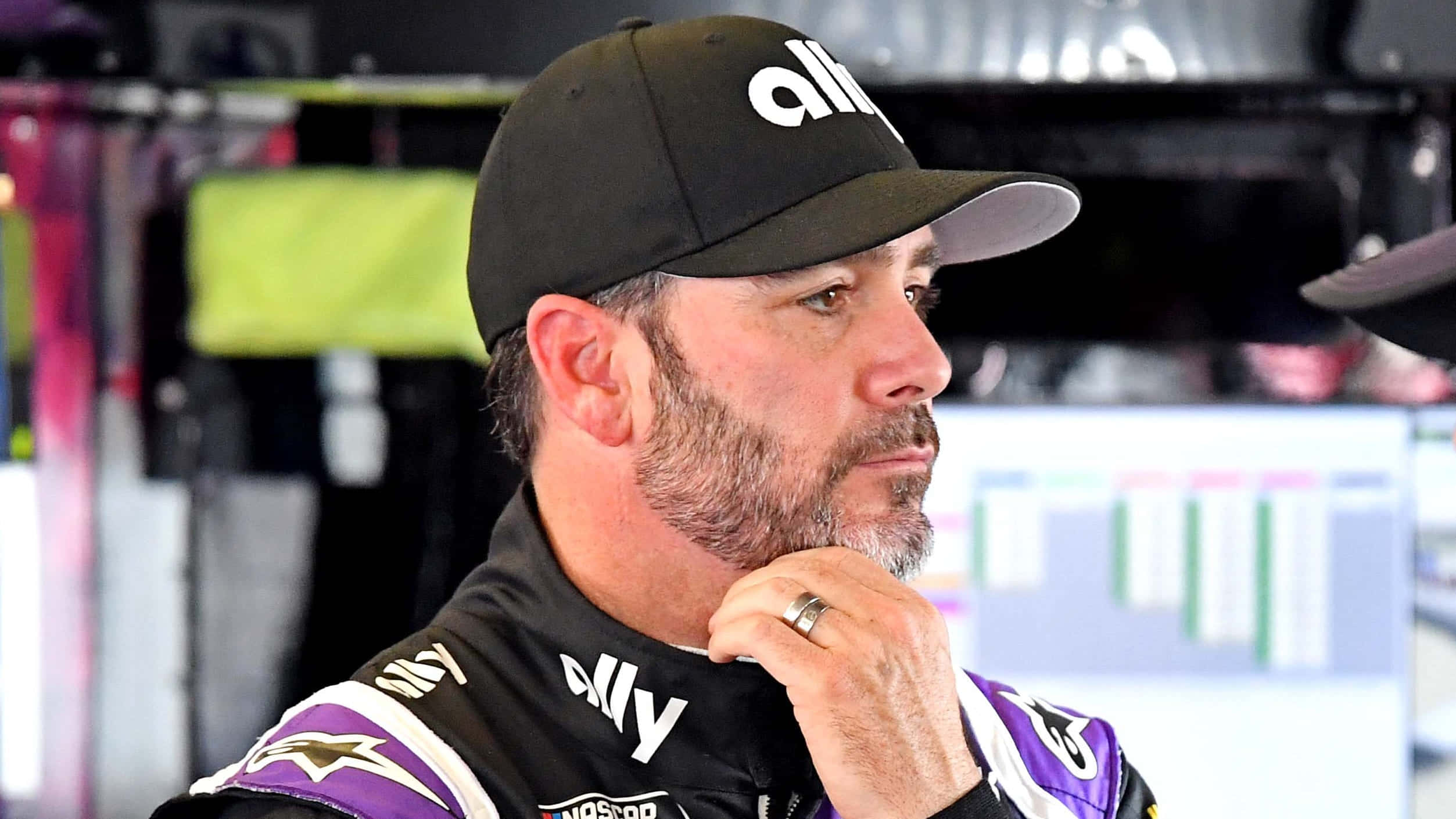 Jimmie Johnson 桌布 壁紙 — 一位身穿黑紫色賽車服和帽子的NASCAR車手，在車庫內托腮沉思。
