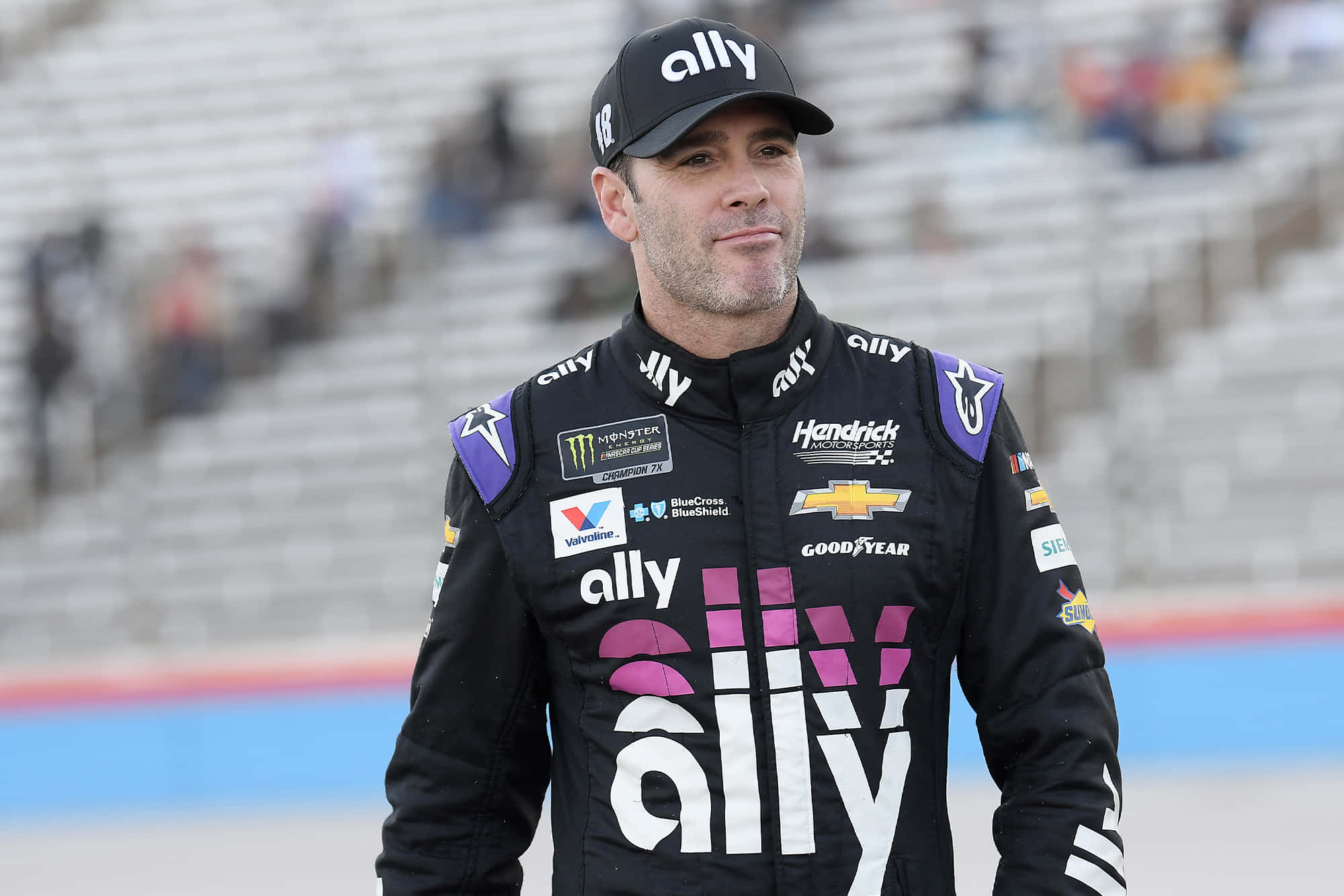 Jimmie Johnson 桌布 壁紙 — 一位身穿黑色Ally賽車服的NASCAR車手自信地站在賽道上，身後是空無一人的看台。