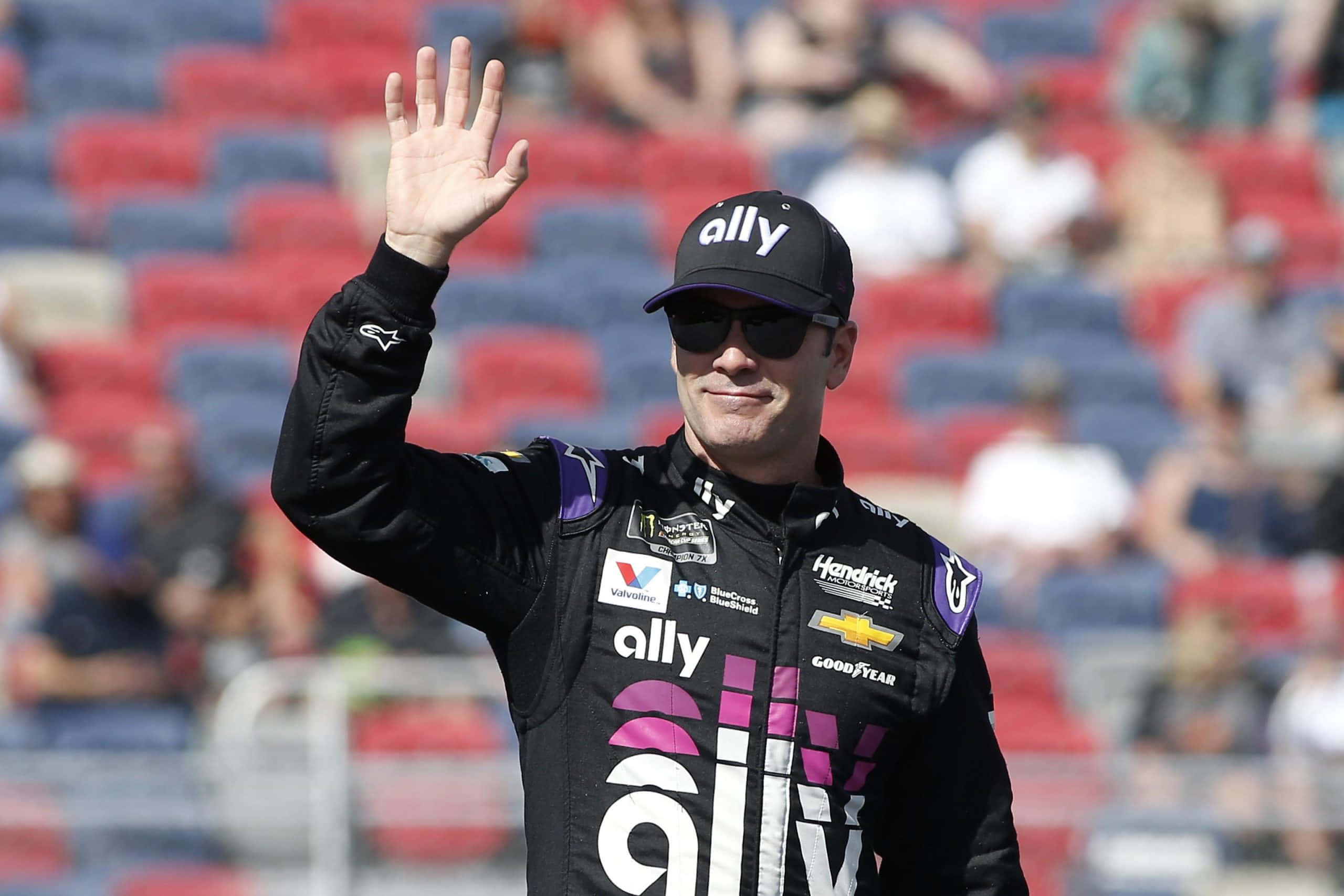 Jimmie Johnson 桌布 壁紙 — 一位身穿黑色Ally賽車服的NASCAR車手在陽光普照、有紅色和藍色座位的體育場向粉絲揮手。