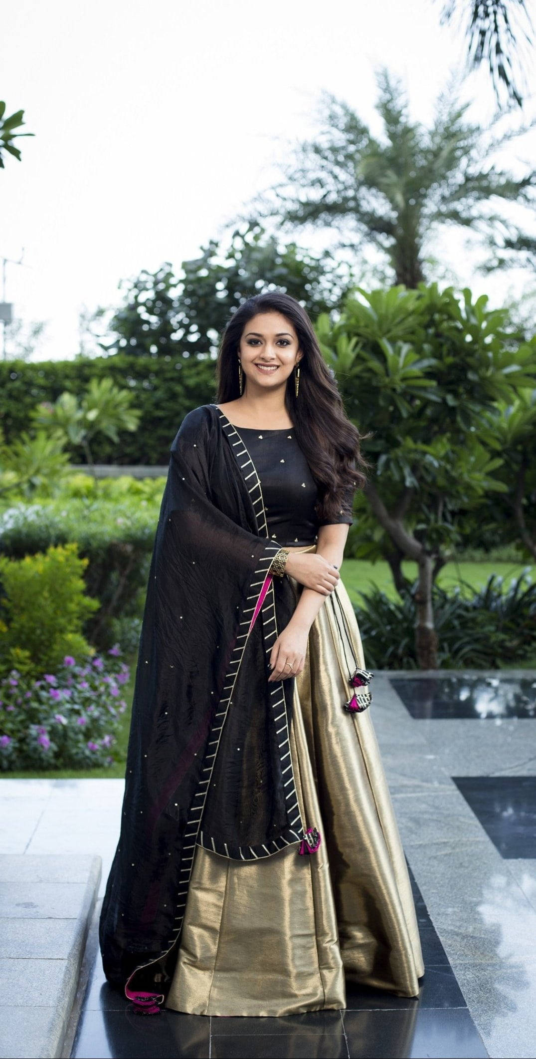 Tamil Actors HD 桌布 壁紙 — 一位身穿黑色和金色傳統服飾的女性在綠意盎然的園林中微笑，園中有棕櫚樹和修剪整齊的樹籬。