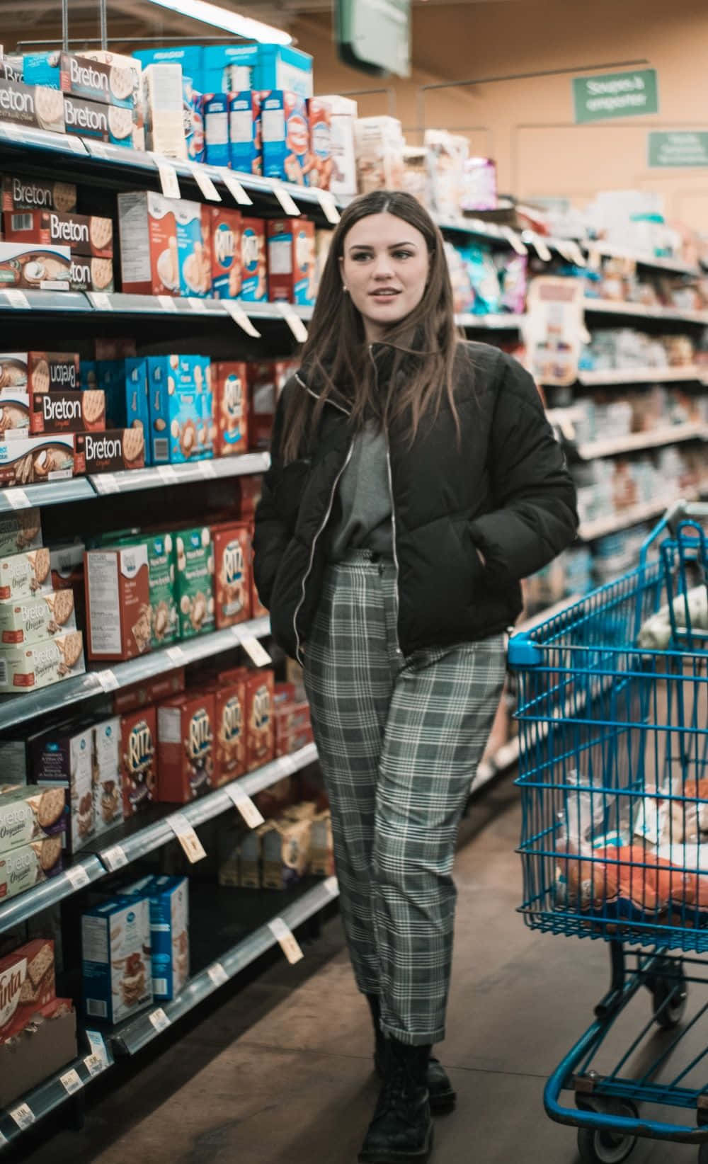 Shopping 桌布 壁紙 — 一位身穿黑色外套和格子褲的年輕女子站在光線充足的雜貨店走道旁，旁邊有一輛藍色購物車。
