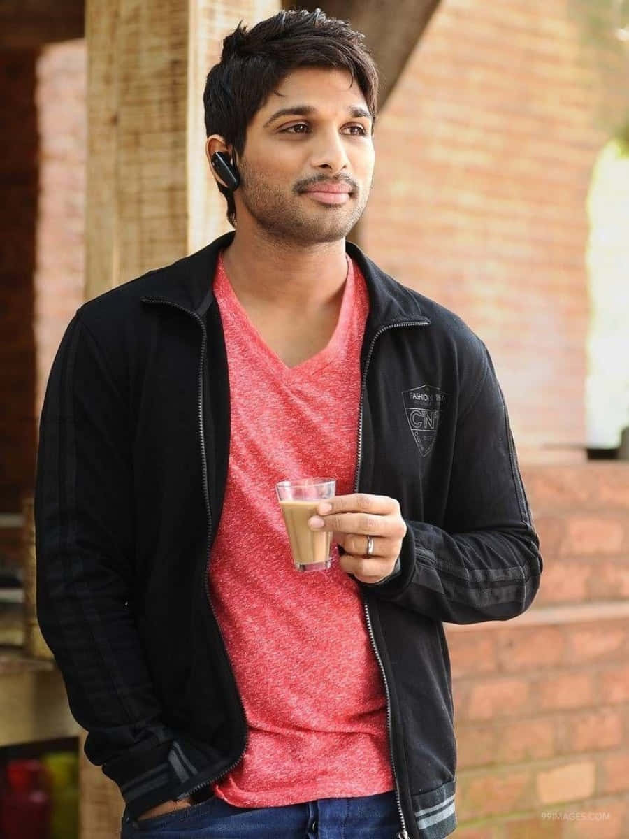 Allu Arjun 桌布 壁紙 — 一位身穿黑色外套和紅色襯衫的年輕男子，在磚牆背景下拿著咖啡杯，戴著無線耳機。