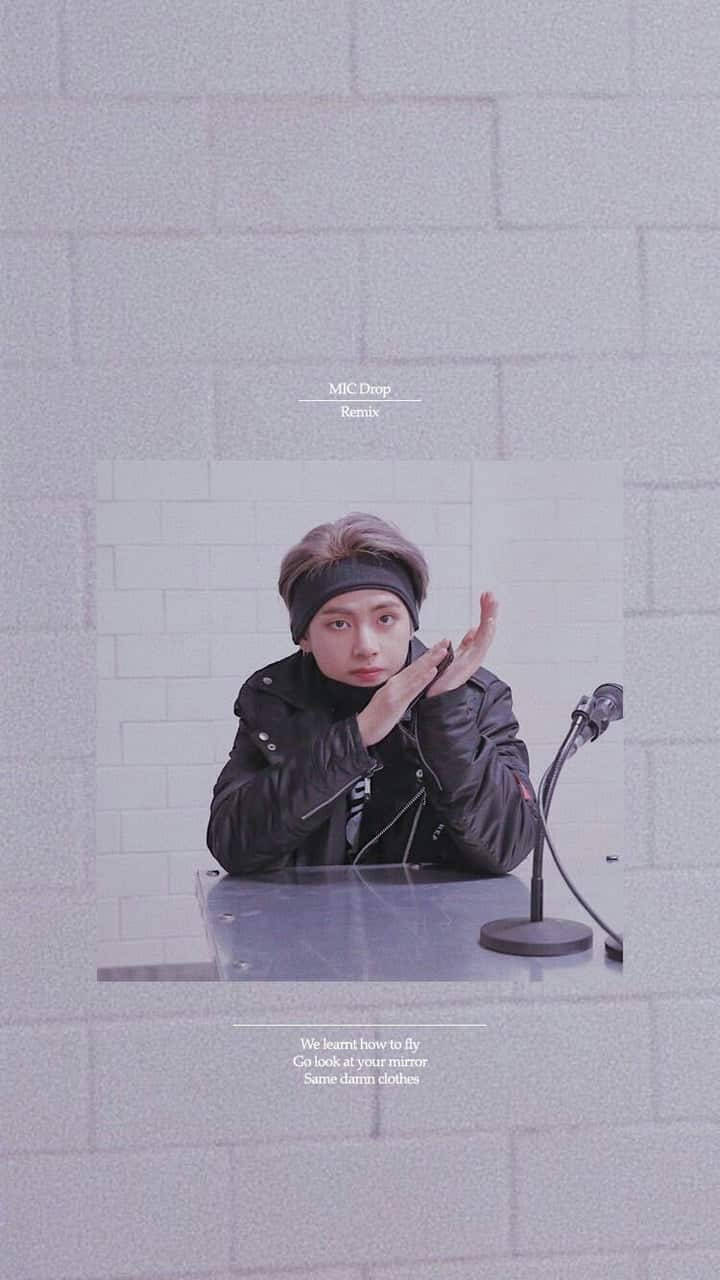 Taehyung《MIC DROP (Remix)》 壁紙 — 一位身穿黑色外套和頭帶的年輕男子坐在桌子旁，桌上放著麥克風，背景是白色磚牆，他正在做手勢。
