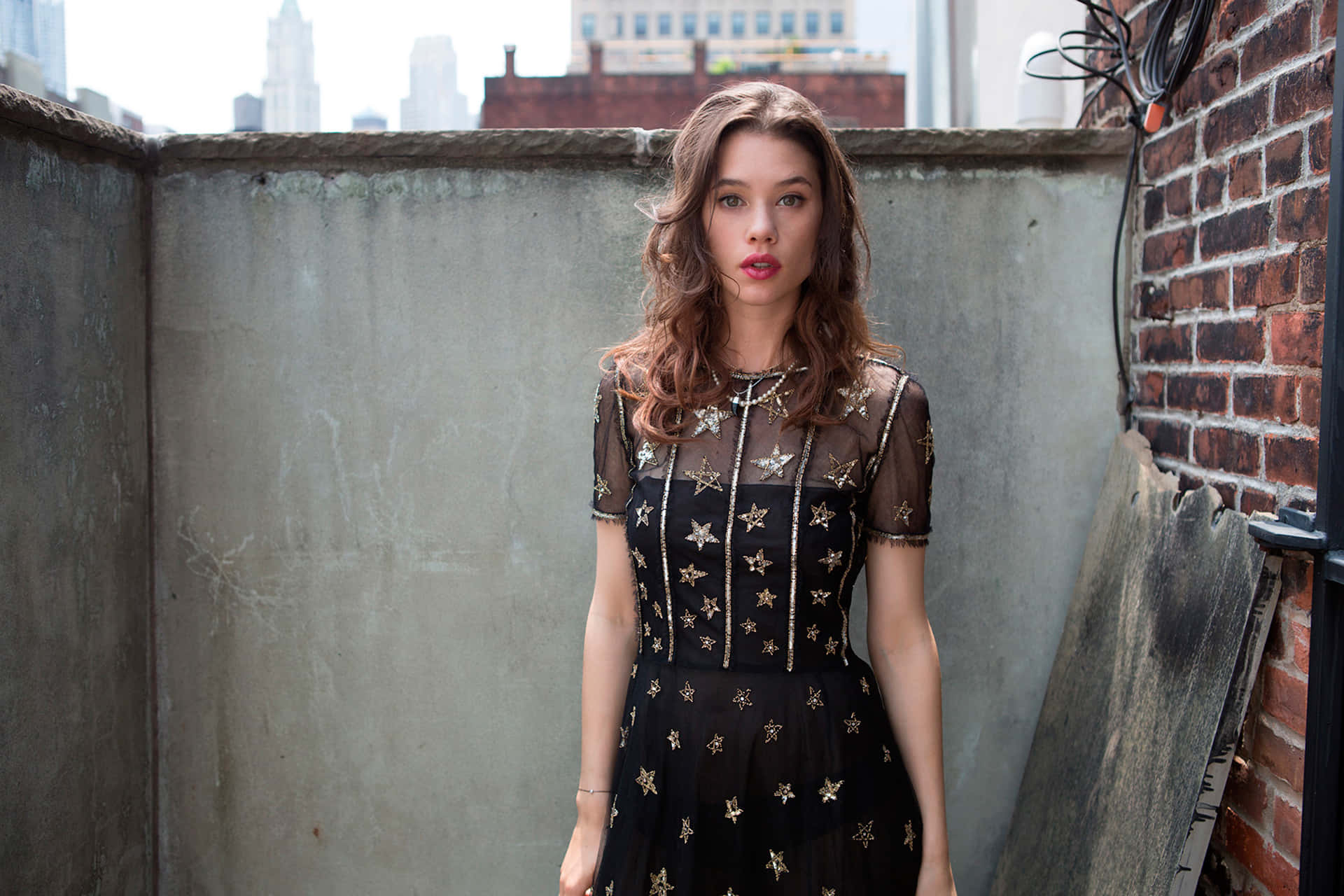 Astrid Berges Frisbey 桌布 壁紙 — 一位身穿黑色星星裝飾連衣裙的年輕女性自信地站在一個粗糙的城市屋頂上，背景是磚牆和城市天際線。