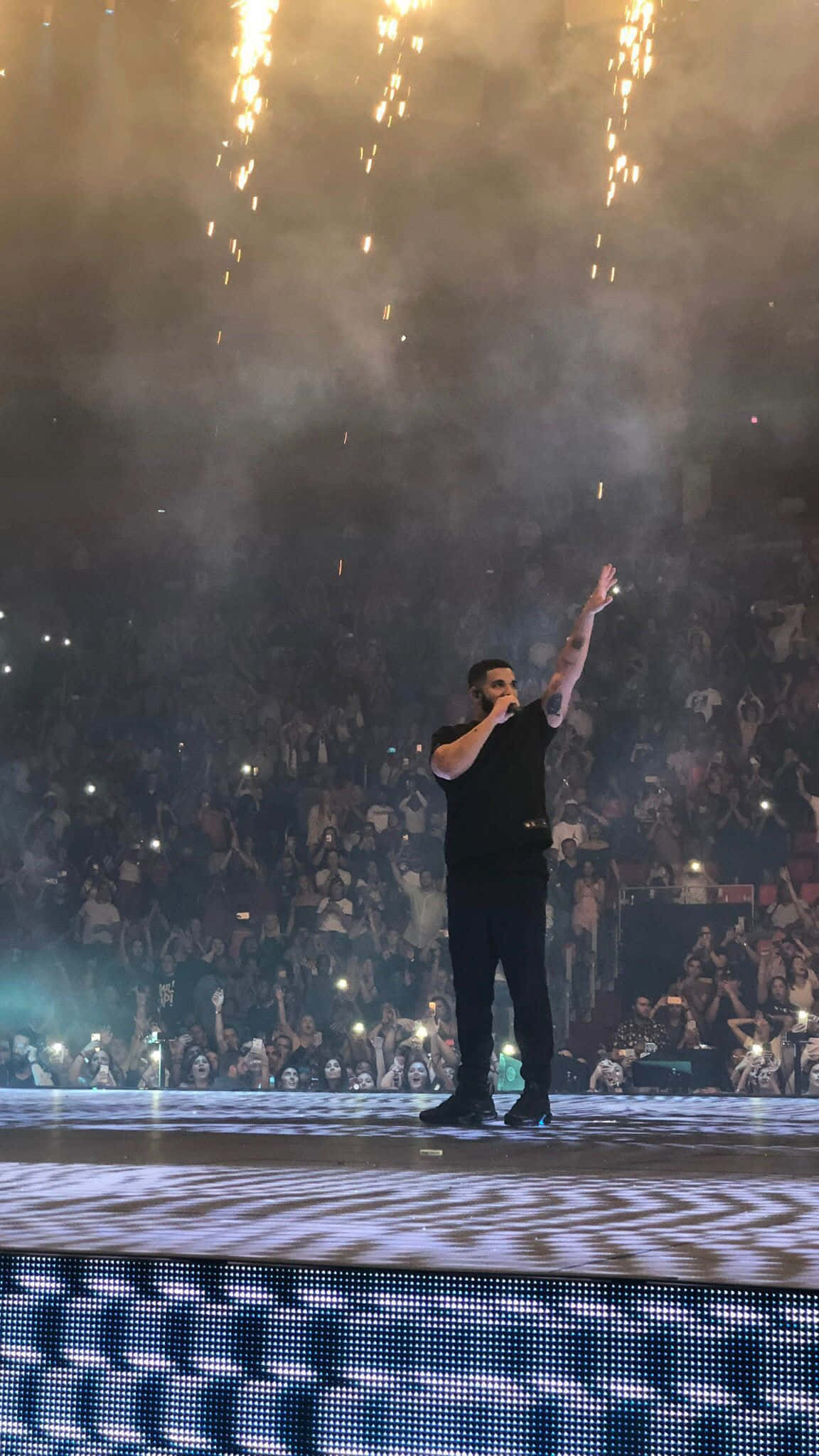 Drake 桌布 壁紙 — 一位身穿黑色服裝的表演者在夜晚的煙火和舞台煙霧中向歡呼的人群揮手。