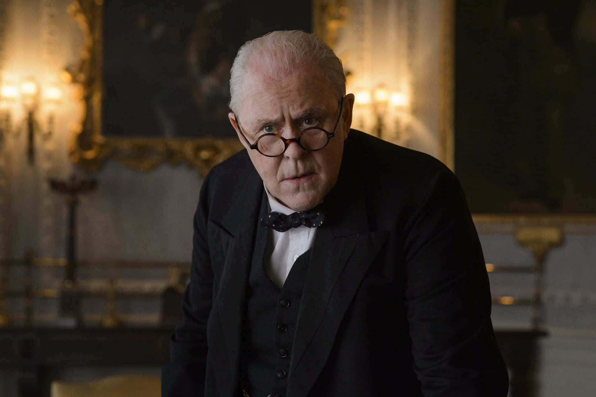 John Lithgow 桌布 壁紙 — 一位身穿黑色正式西裝、戴著圓框眼鏡的長者，在一個光線昏暗、裝飾華麗且帶有金色點綴的房間裡專注地凝視。