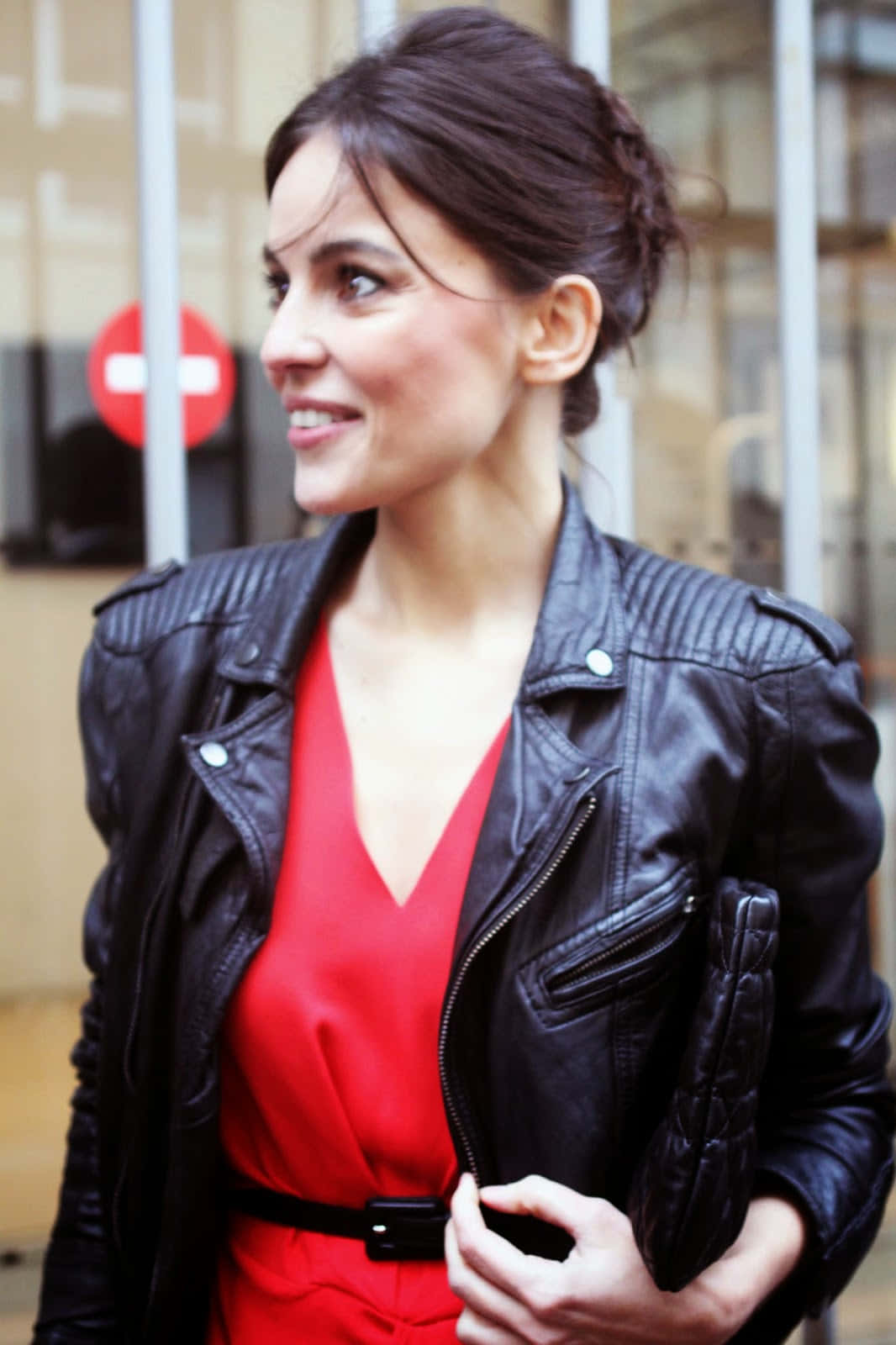 Elena Anaya 桌布 壁紙 — 一位身穿黑色皮褸和紅色連身裙的女性，在玻璃建築附近戶外手持手拿包微笑。