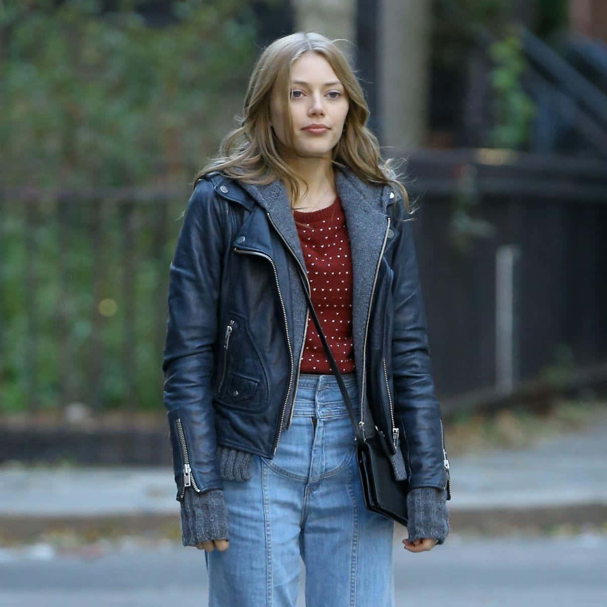 Grace Van Dien 桌布 壁紙 — 一位身穿黑色皮褸和藍色牛仔褲的年輕女性自信地走在城市街道上，周圍散落著秋天的落葉。