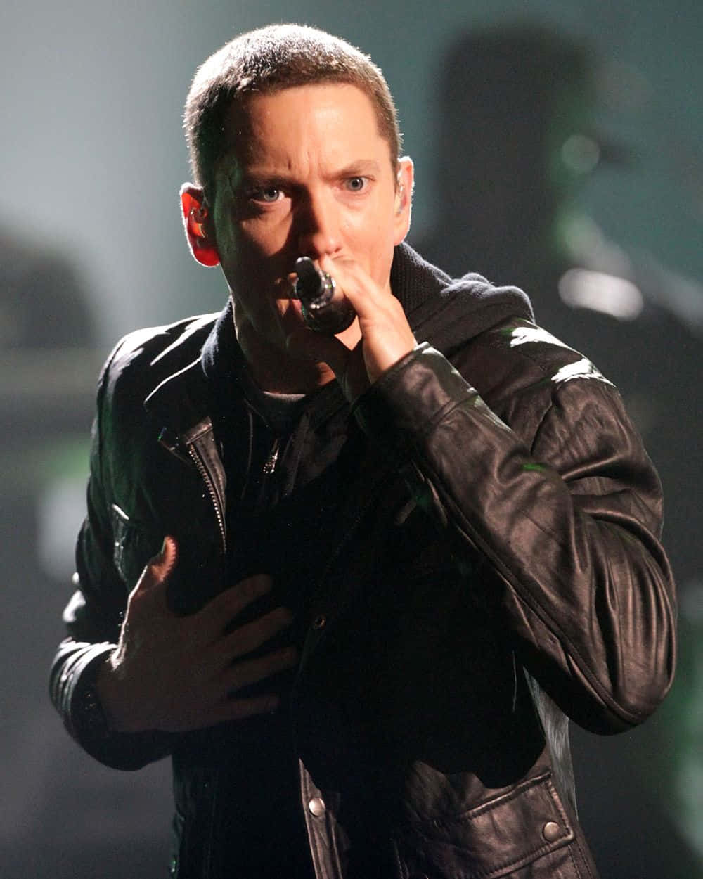 Eminem 桌布 壁紙 — 一位身穿黑色皮褸的男表演者在舞台燈光下熱情地對著咪高峰唱歌。