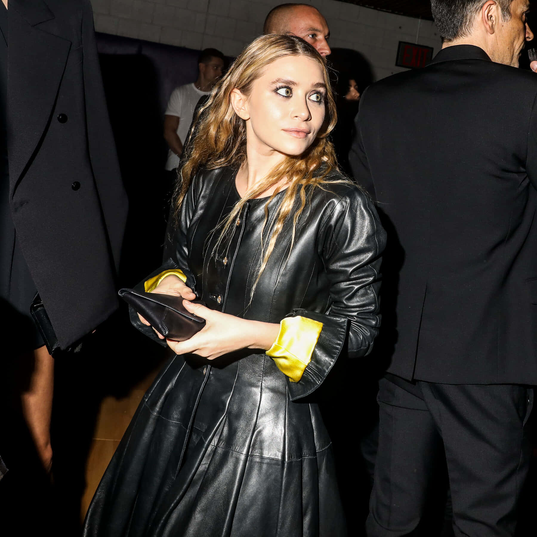 ashley olsen 桌布 壁紙 — 一位身穿黑色皮革連衣裙、帶有黃色緞面袖口的女士，在光線昏暗的室內環境中手持手拿包，直視鏡頭。