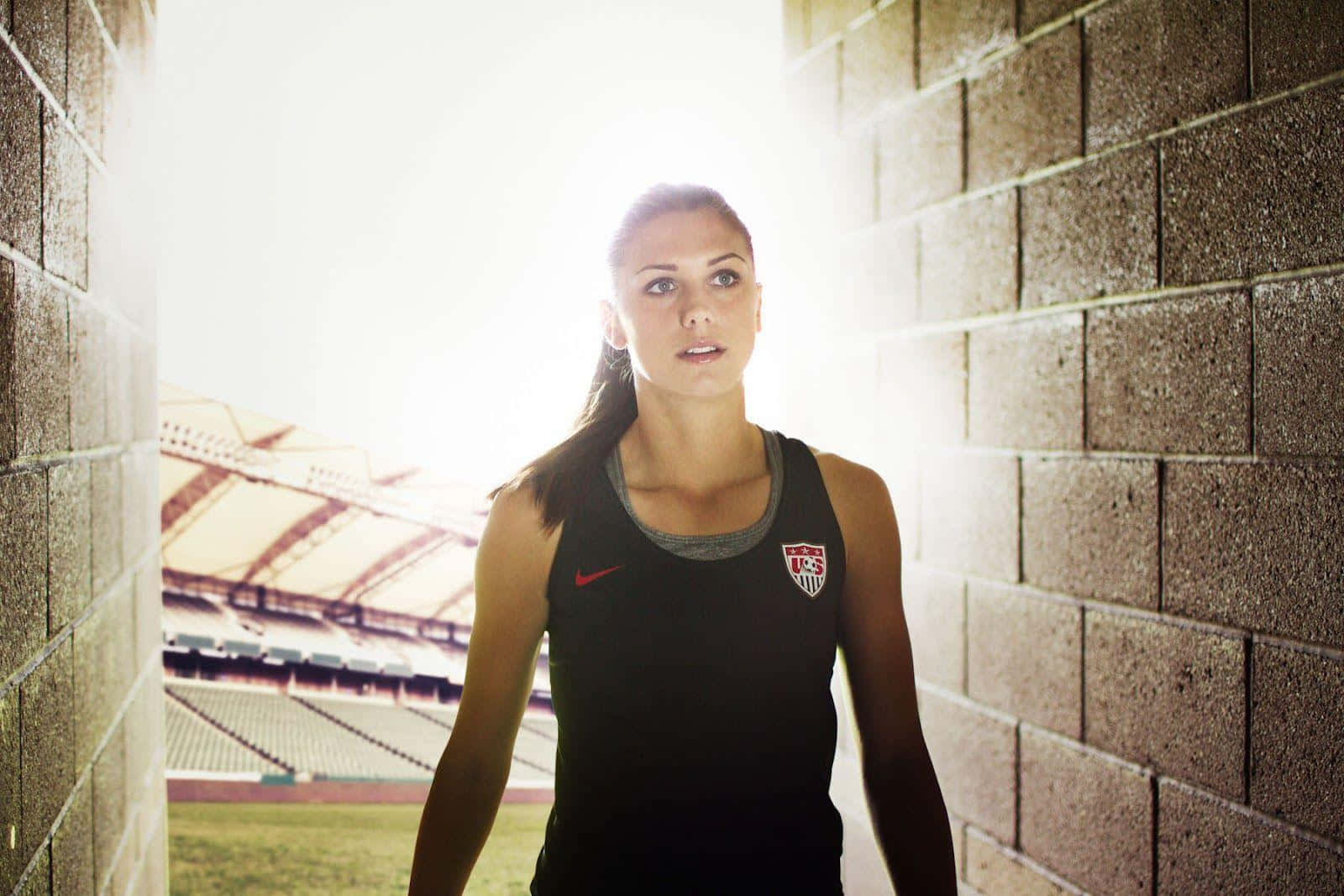 Alex Morgan 桌布 壁紙 — 一位身穿黑色美國足球球衣的女運動員，站在陽光普照的體育場隧道中，堅定地向前望。
