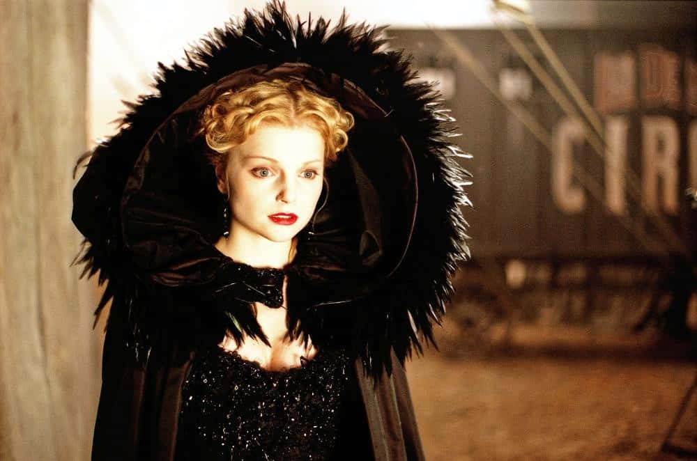 Izabella Miko 桌布 壁紙 — 一位身穿黑色羽毛斗篷的女性，在昏暗的馬戲團背景下，以紅唇和銳利的目光從容站立。