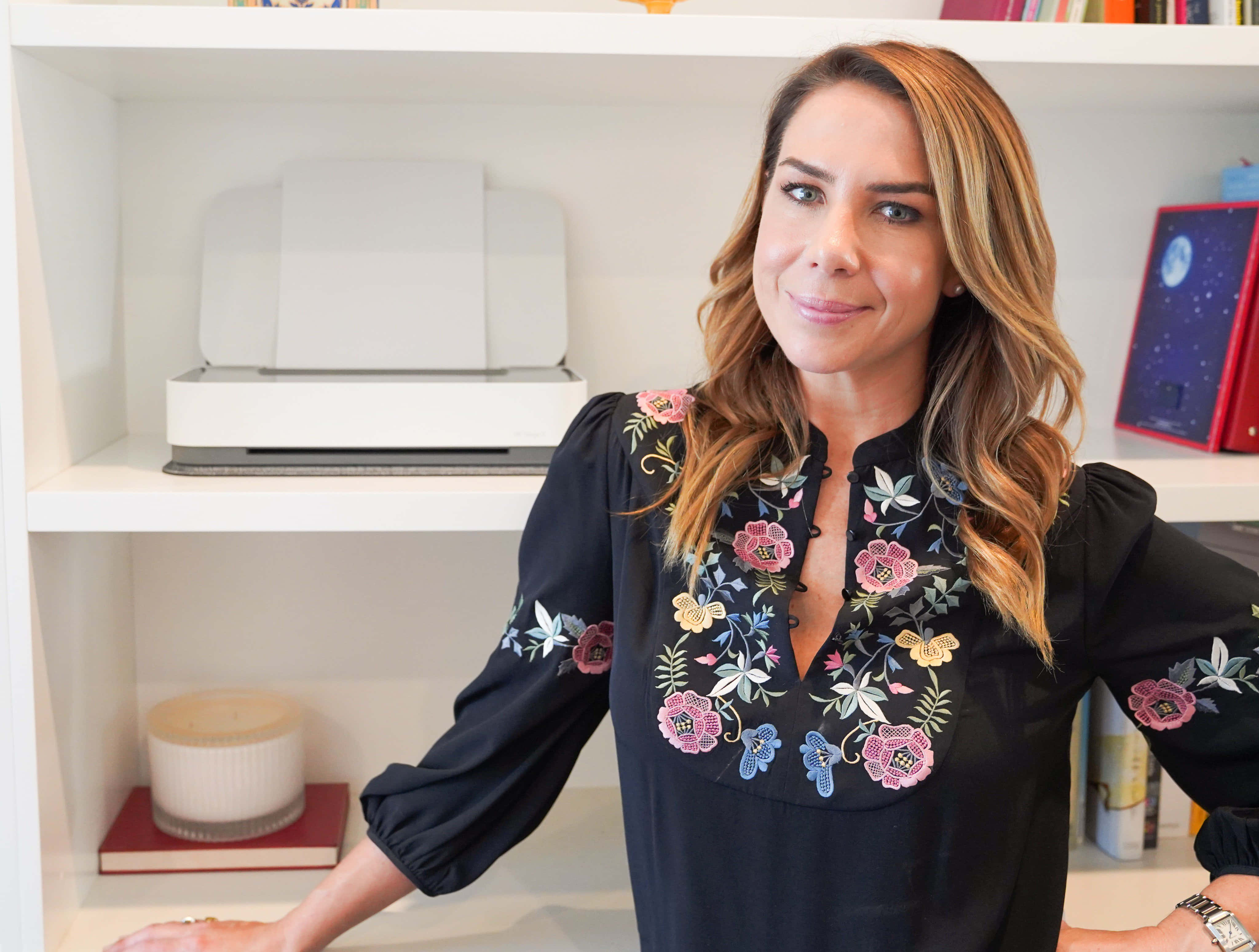 Kate Ritchie 桌布 壁紙 — 一位身穿黑色花卉刺繡襯衫的女性微笑著靠在一個放有打印機和蠟燭的白色書架上。