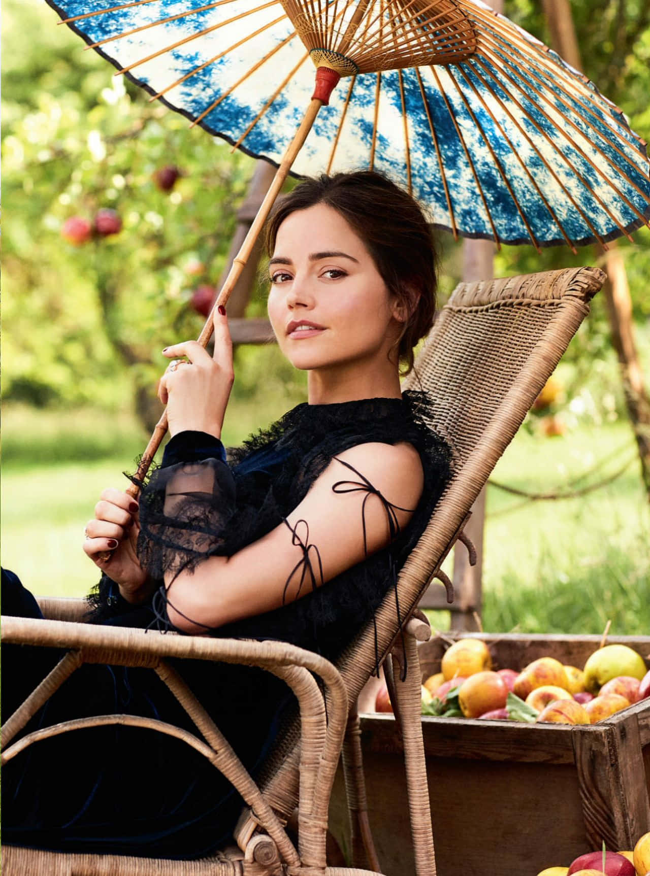 Jenna Coleman 桌布 壁紙 — 一位身穿黑色蕾絲裙的女子在陽光明媚的果園裡，坐在藍色圖案陽傘下的柳條椅上，旁邊放著一箱蘋果，悠閒地休息。