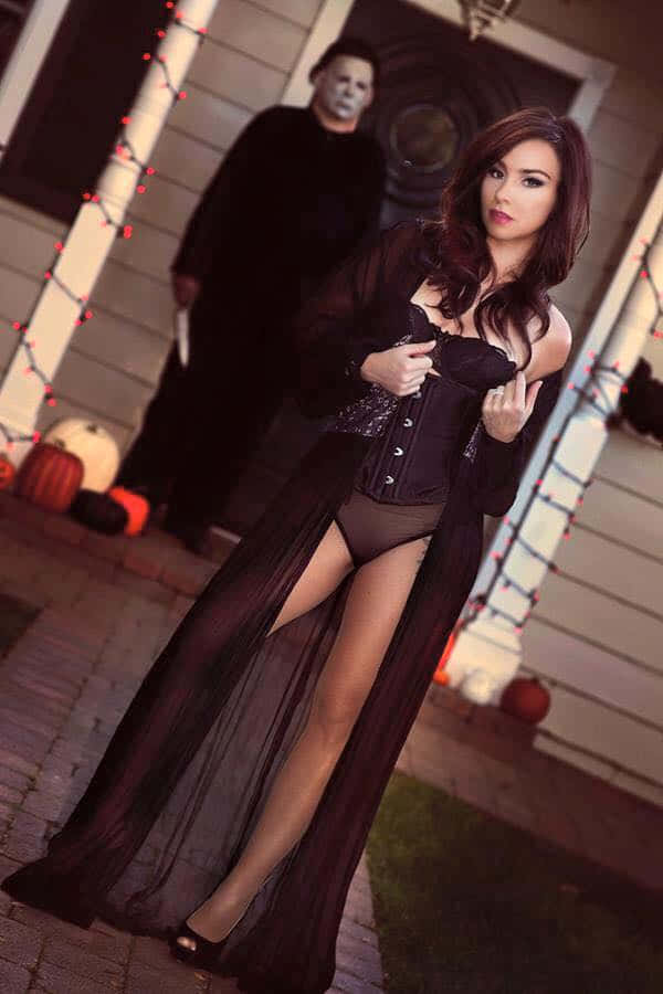 Danielle Harris 桌布 壁紙 — 一位身穿黑色薄紗長裙的女性在門廊上自信地擺姿勢，而一個戴面具的身影在萬聖節裝飾中潛伏在她身後。