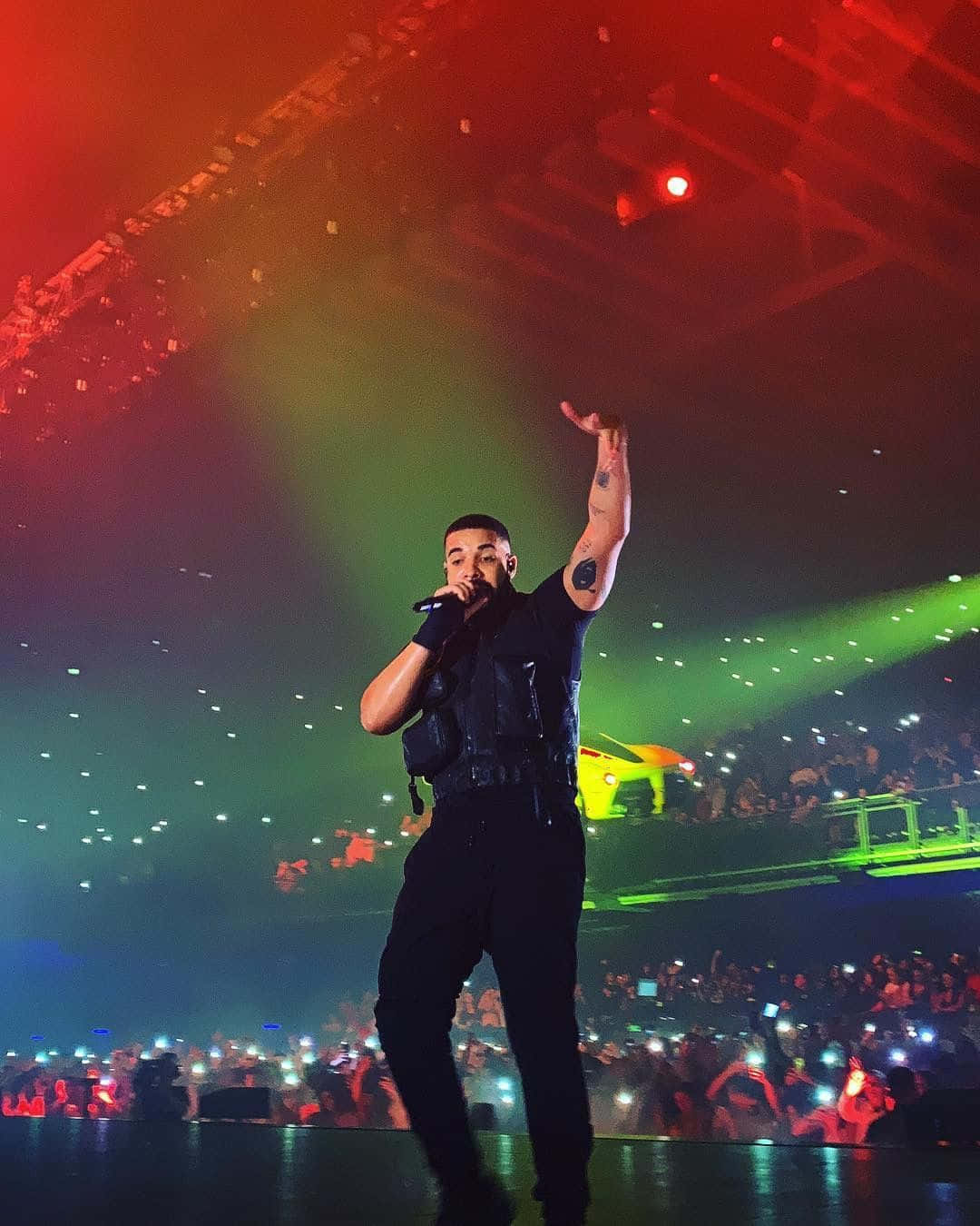 Drake 桌布 壁紙 — 一位身穿黑色衣服的男性表演者在燈光璀璨的舞台上，對著咪高峰唱歌，舞台上有紅色和綠色的射燈，觀眾舉起手機。