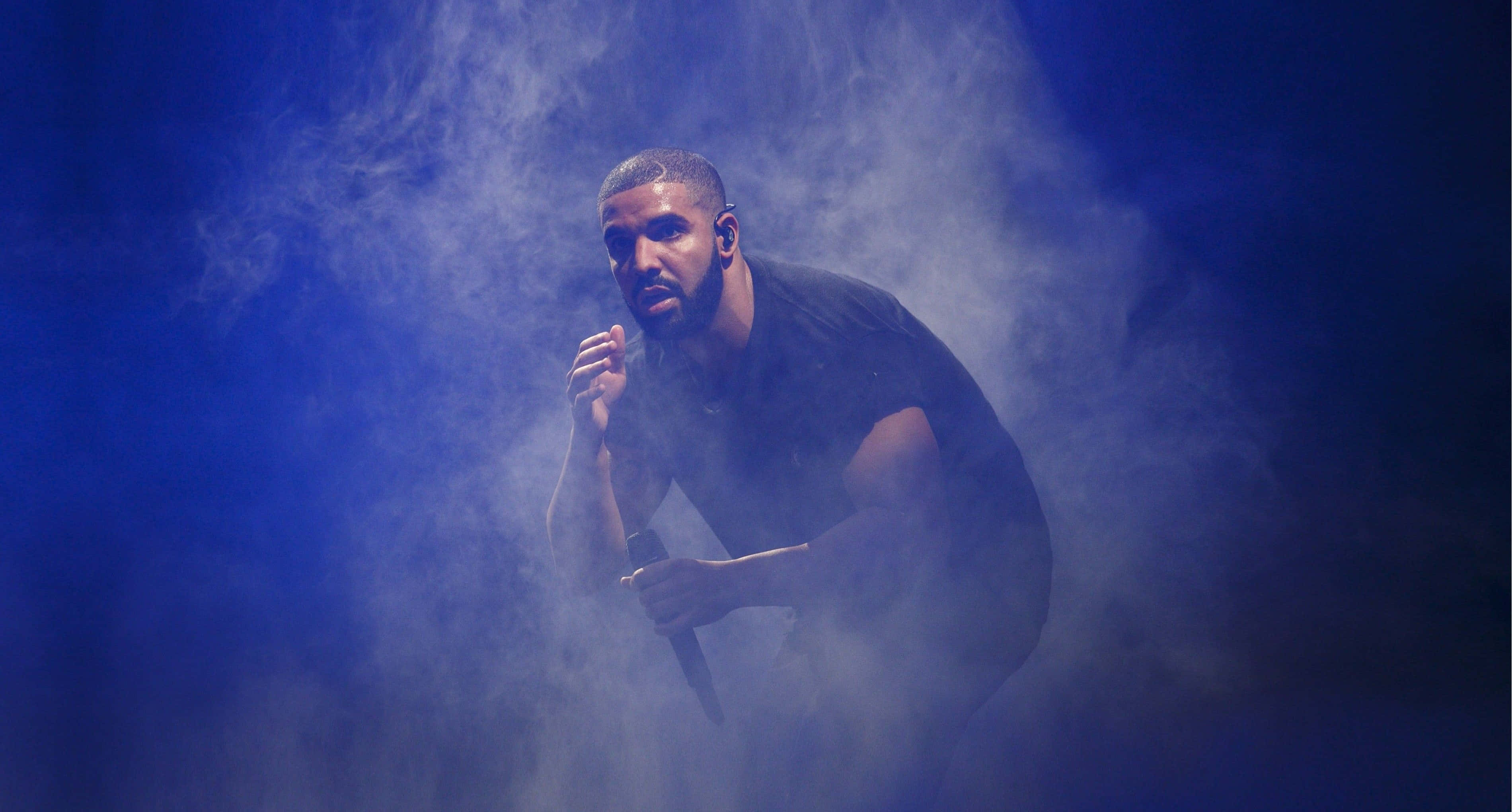 Drake 桌布 壁紙 — 一位身穿黑色襯衫的男性表演者，在煙霧瀰漫、藍色燈光的舞台環境中蹲下，手持麥克風。